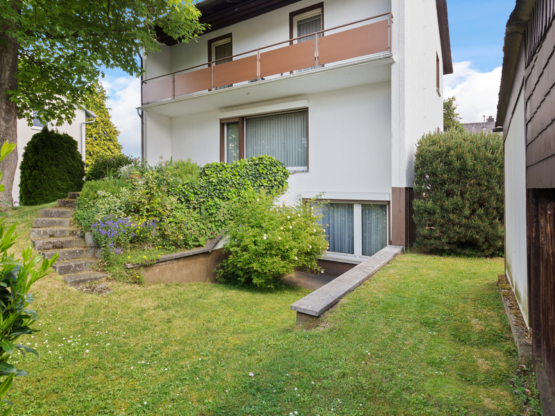 Garten mit Rasen, Sträuchern und Treppe neben einem weißen Wohnhaus mit Balkon.