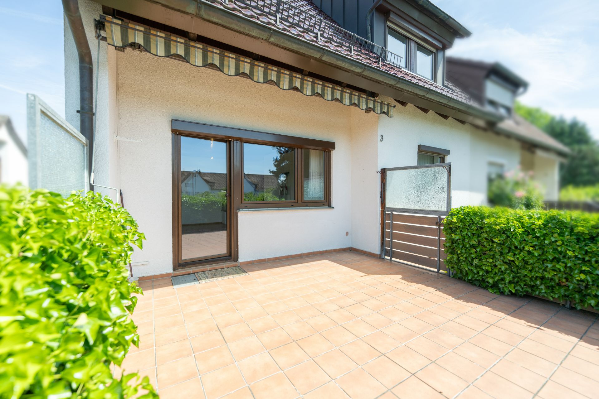 Geflieste Terrasse mit Markise, Glastrennwand und Hecken vor einem Wohnhaus.