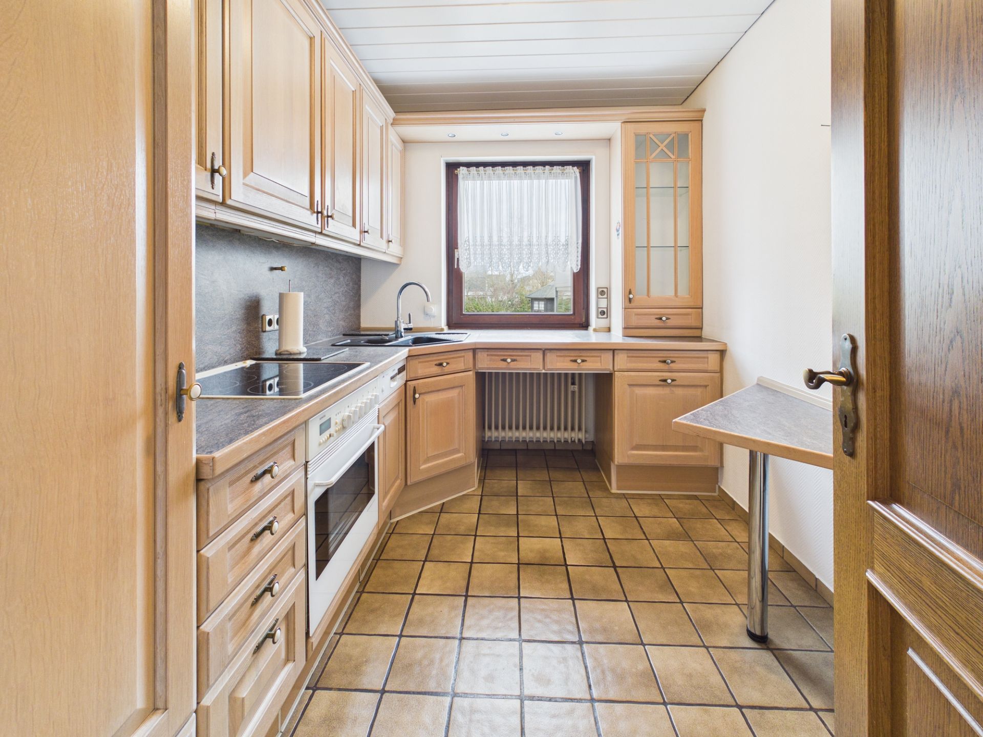 Helle Einbauküche mit Holzfronten, Fenster, Herd, Spüle und Fliesboden.