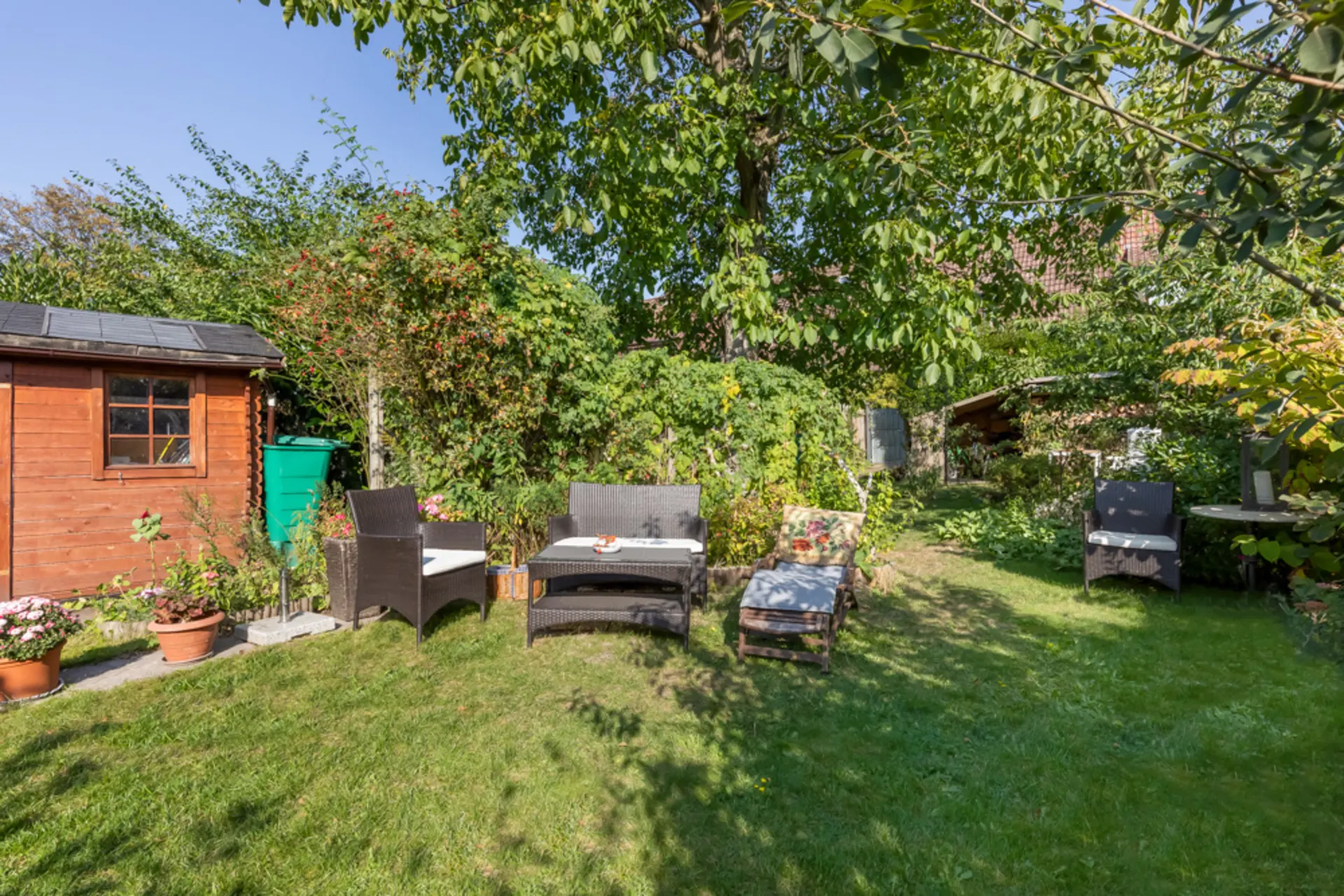 Grüner Garten mit Gartenmöbeln, Holzhaus und vielen Pflanzen bei sonnigem Wetter.