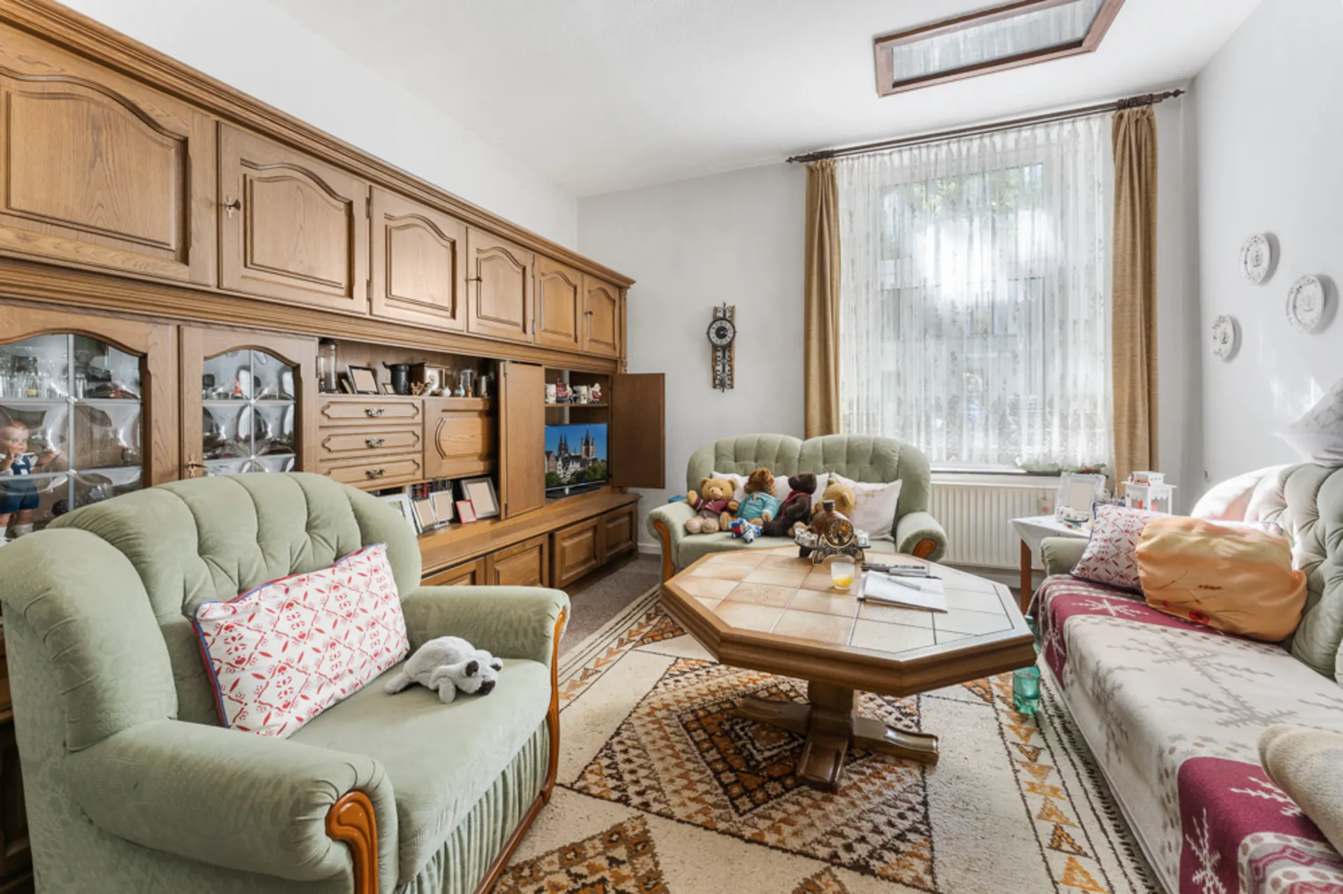 Wohnzimmer mit grünen Polstermöbeln, Holzwandschrank, Couchtisch und großen Fenstern.