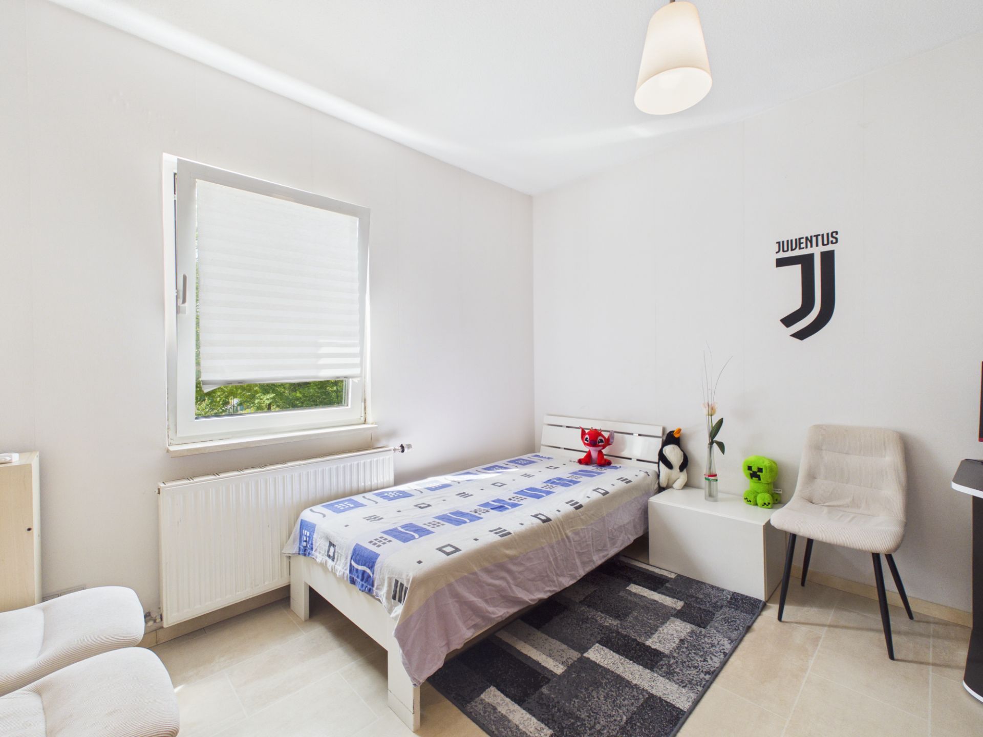 Helles Schlafzimmer mit Einzelbett, Stuhl, Nachttisch und Fenster mit Rollos.