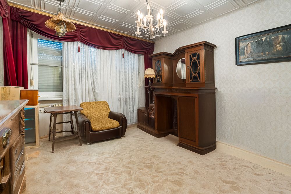 Wohnzimmer mit Teppichboden, Sessel, Holzmöbeln und Vorhängen vor großen Fenstern.