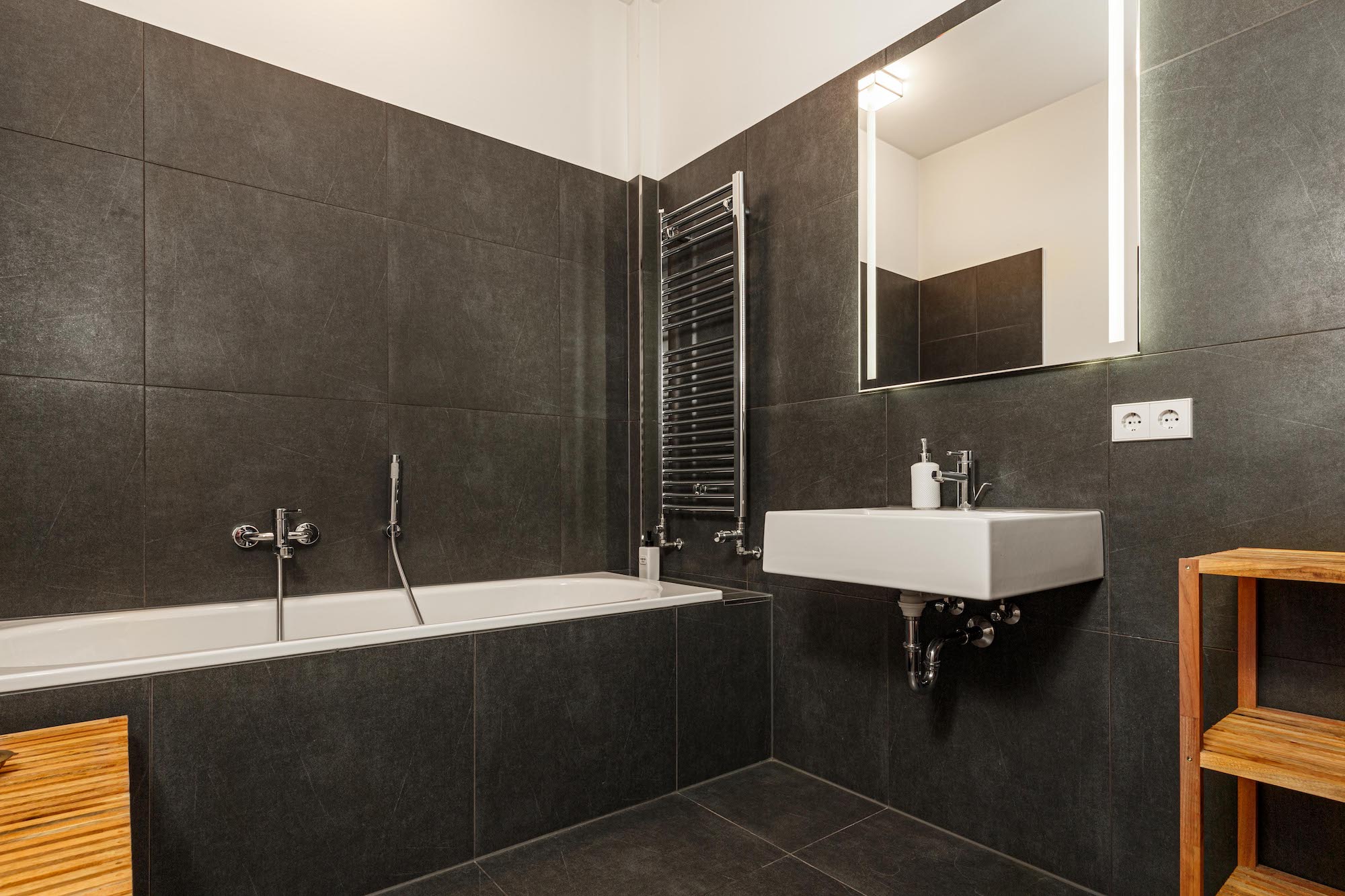 Modernes Badezimmer mit dunklen Fliesen, Badewanne, Waschbecken und beleuchtetem Spiegel.