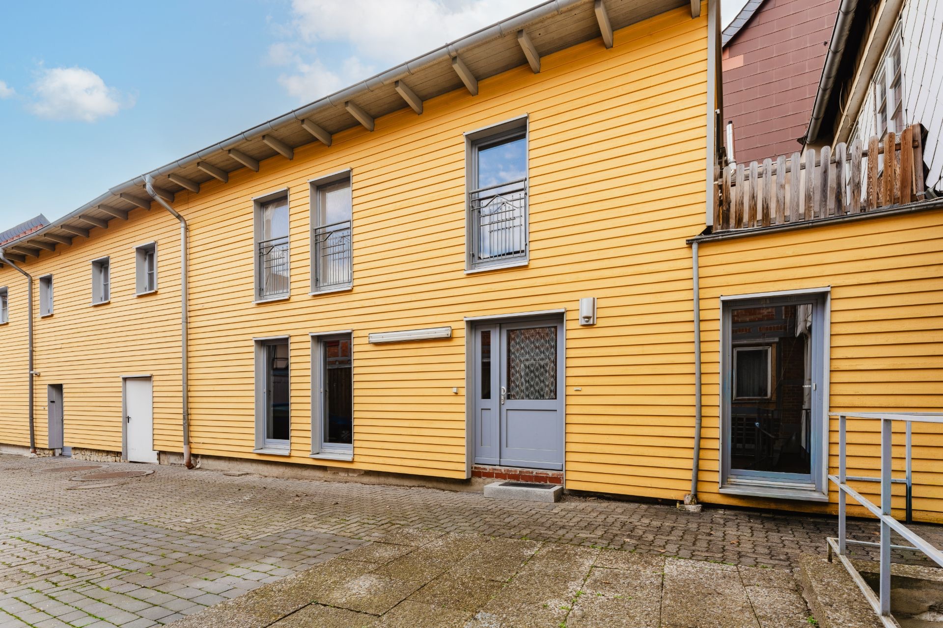 Moderne gelbe Hausfassade mit mehreren Fenstern und Türen, gepflasterter Hof im Vordergrund.