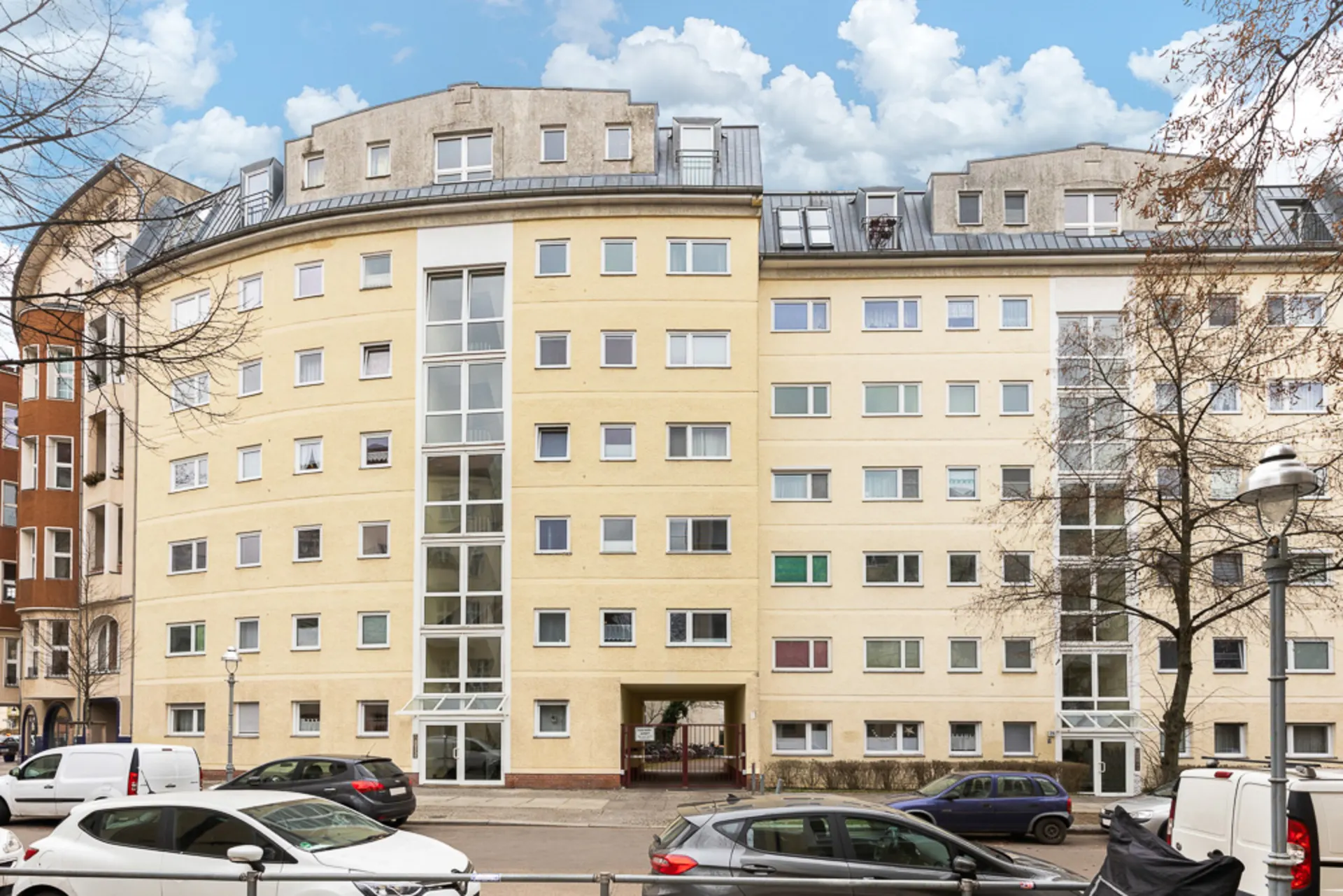 Mehrstöckiges, gelbes Wohngebäude mit vielen Fenstern und Glas-Eingangsbereichen, davor parkende Autos.