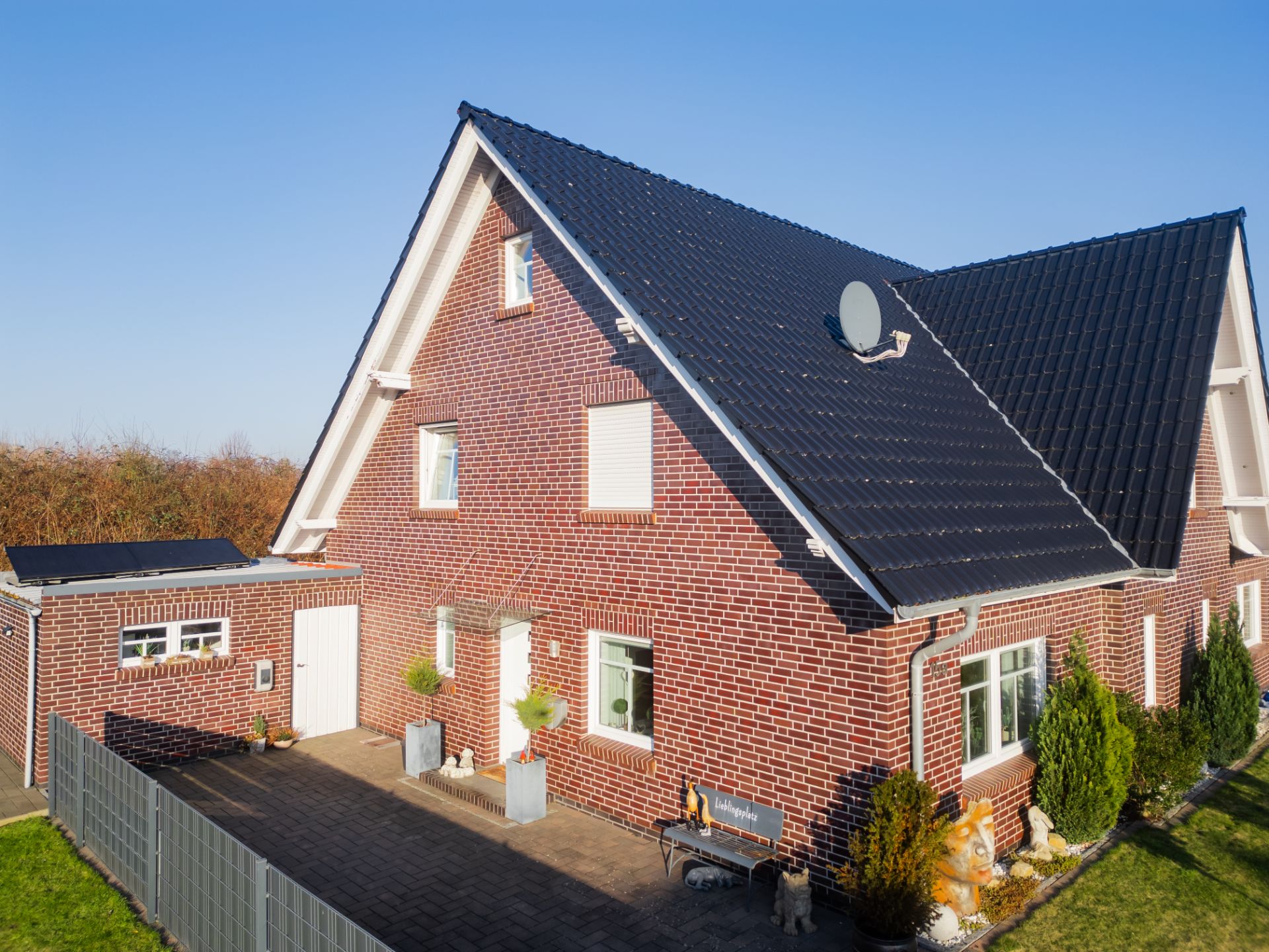 Backsteinhaus mit schwarzem Dach, Vorgarten und Terrasse bei sonnigem Wetter.