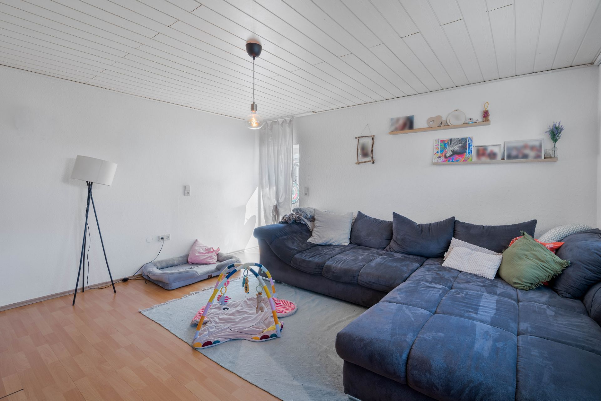 Wohnzimmer mit dunkelblauem Sofa, Stehlampe, Babyspieldecke und Wandregalen.