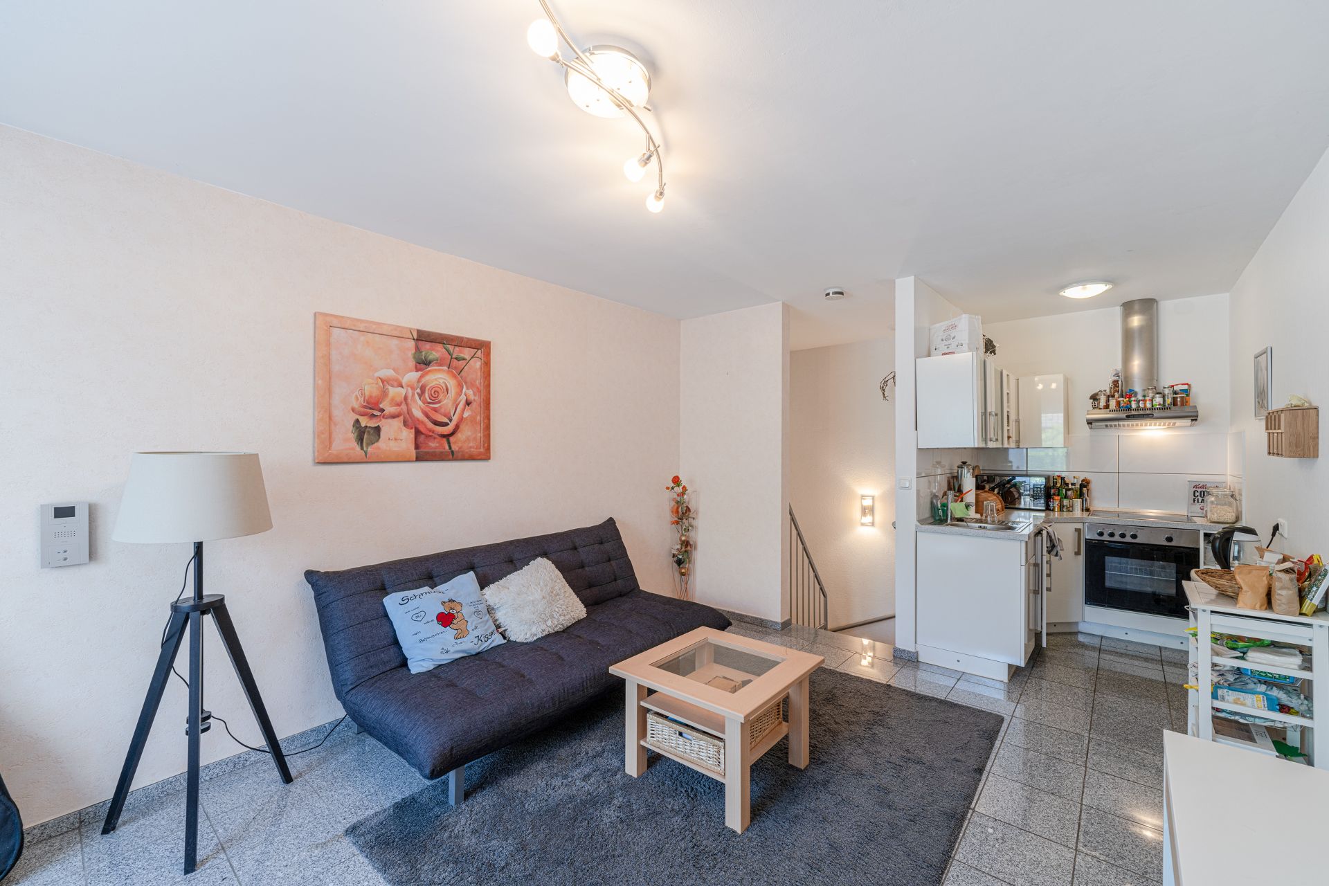 Wohnzimmer mit schwarzem Sofa, Stehlampe, Couchtisch und offener Küche, Bild mit Rosen an der Wand.