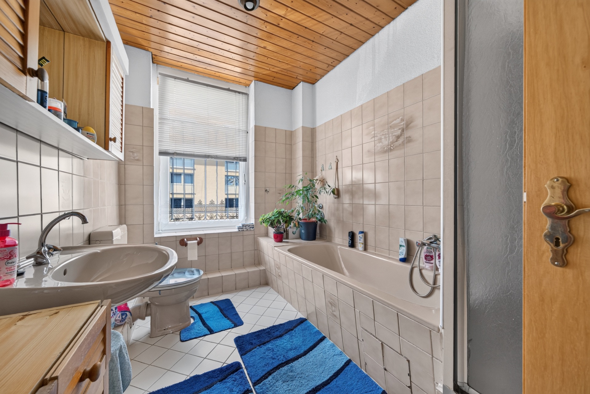 Badezimmer mit Badewanne, Waschbecken, WC und blauen Teppichen, großes Fenster und Pflanzen auf dem Wannenrand.