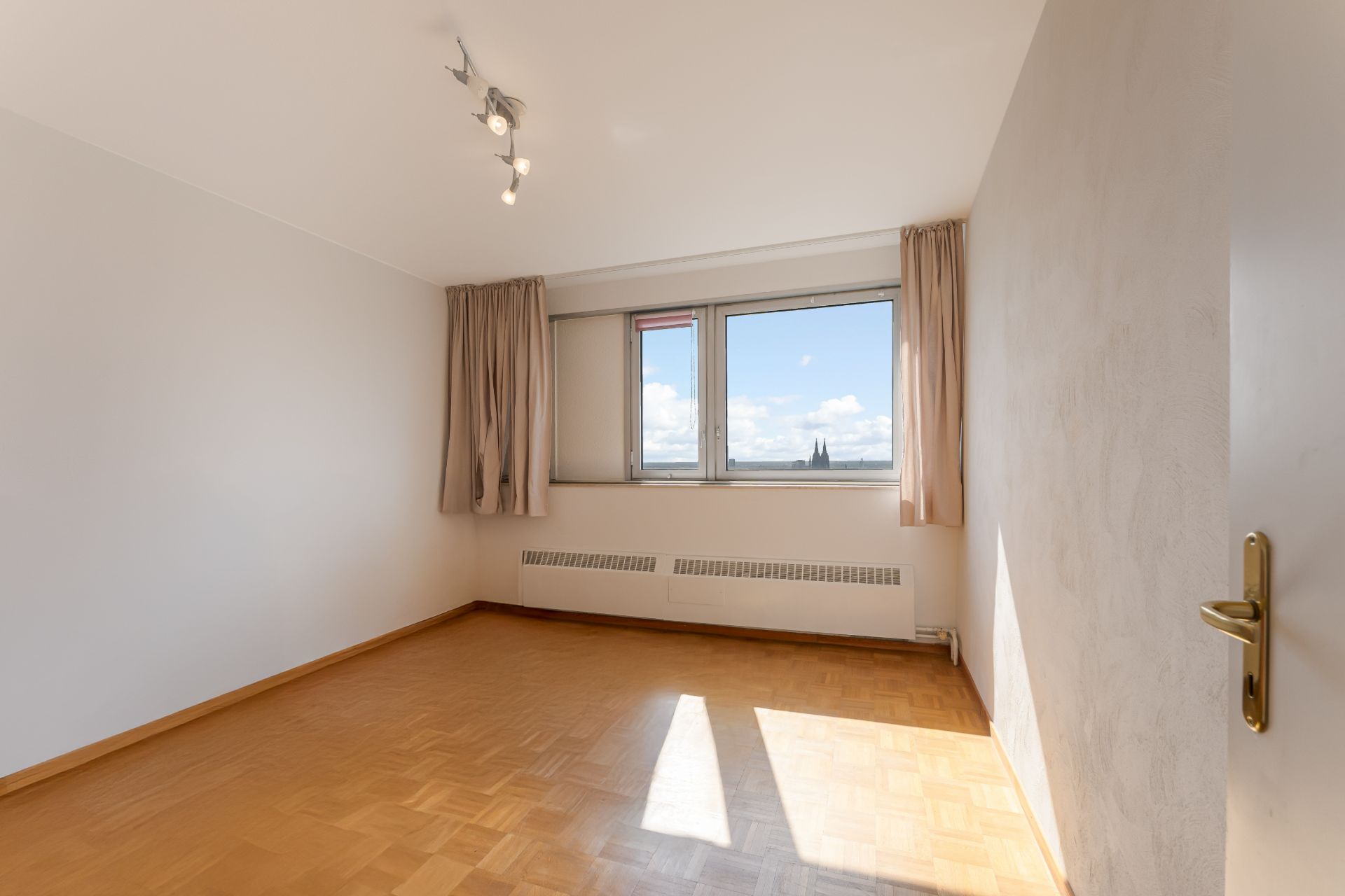 Helles, leeres Zimmer mit Parkettboden, Fenster und beige Vorhängen.