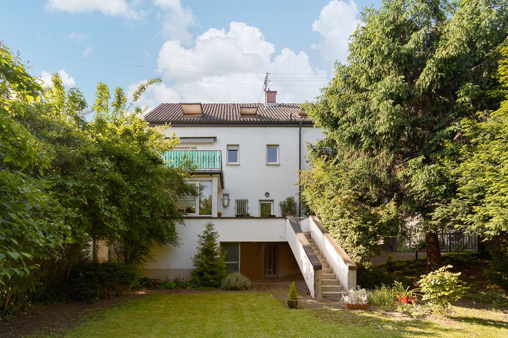 Rückseite eines weißen Wohnhauses mit Balkon, Außentreppe und Garten, umgeben von Bäumen.