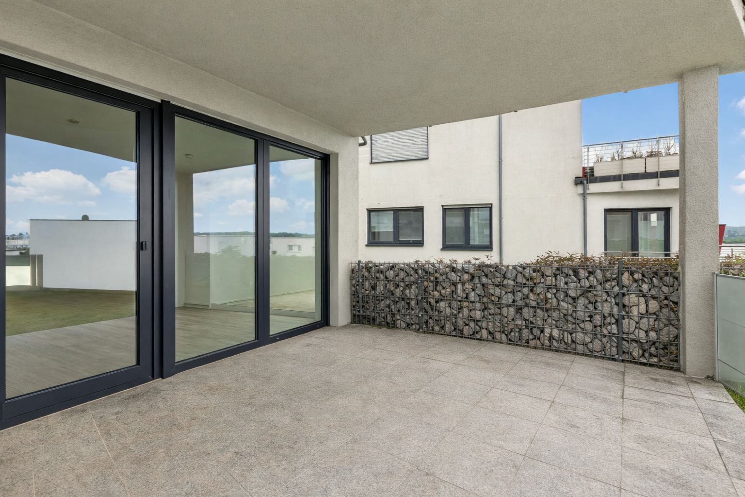 Überdachte Terrasse mit Fliesenboden, Glasgeländer und Steinmauer, Blick auf benachbartes Gebäude.
