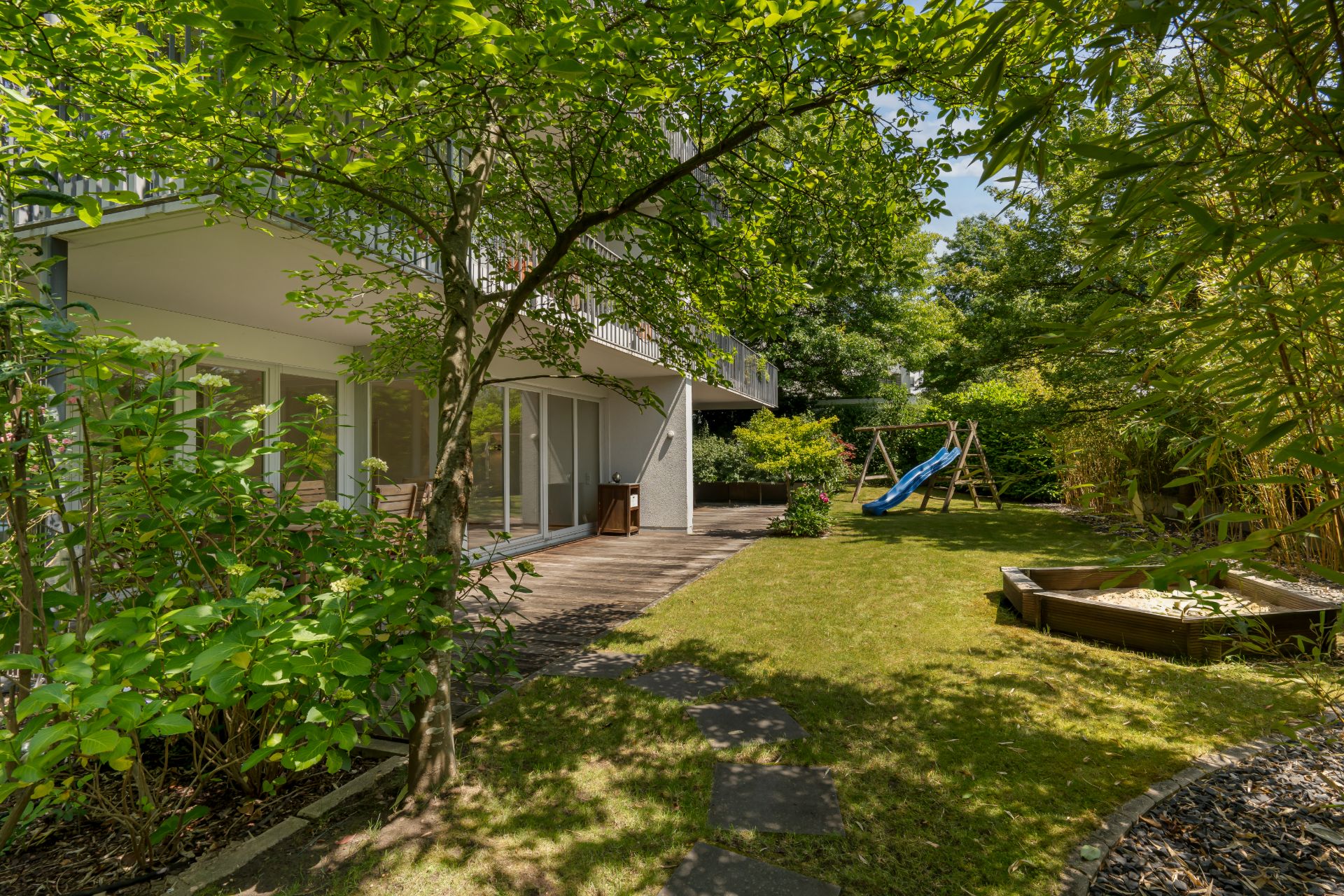 Garten mit Terrasse, Rutsche, Sandkasten und viel Grün an einem Wohnhaus.