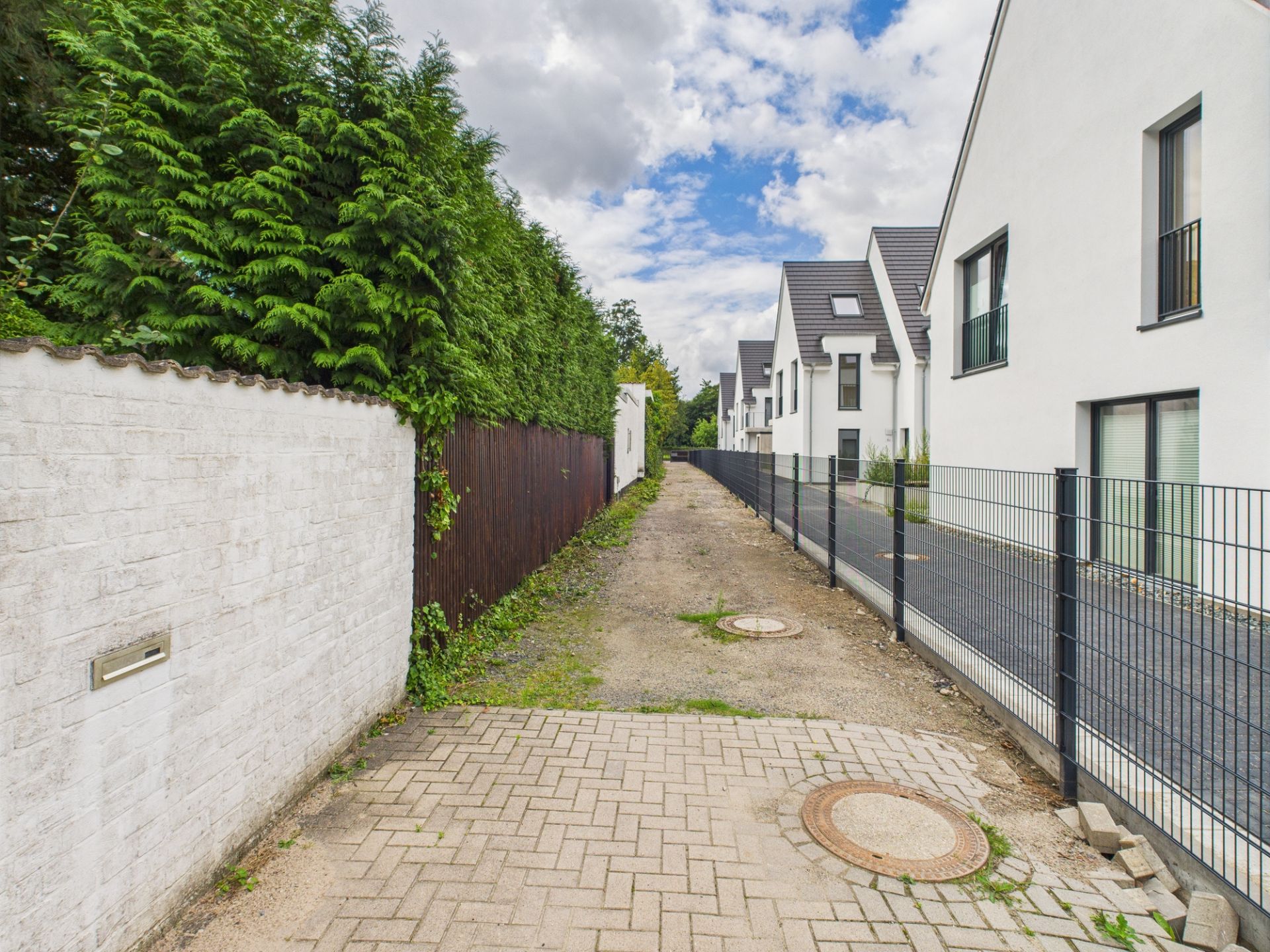 Gepflasterter Weg mit Gullydeckeln neben weißen Wohnhäusern und Hecke.