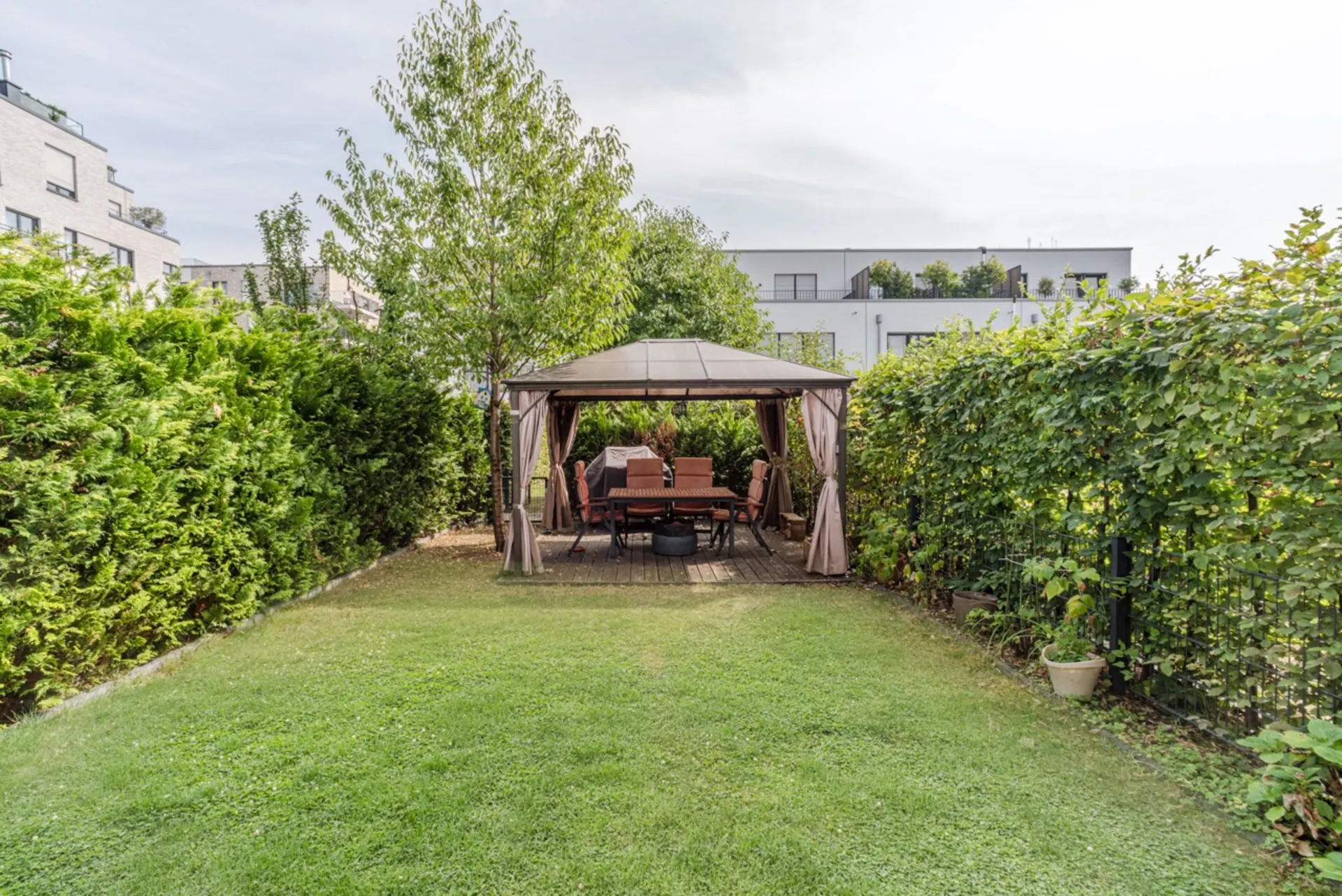 Garten mit Pavillon, darunter Sitzgruppe und Tisch, umgeben von Hecken und Bäumen.