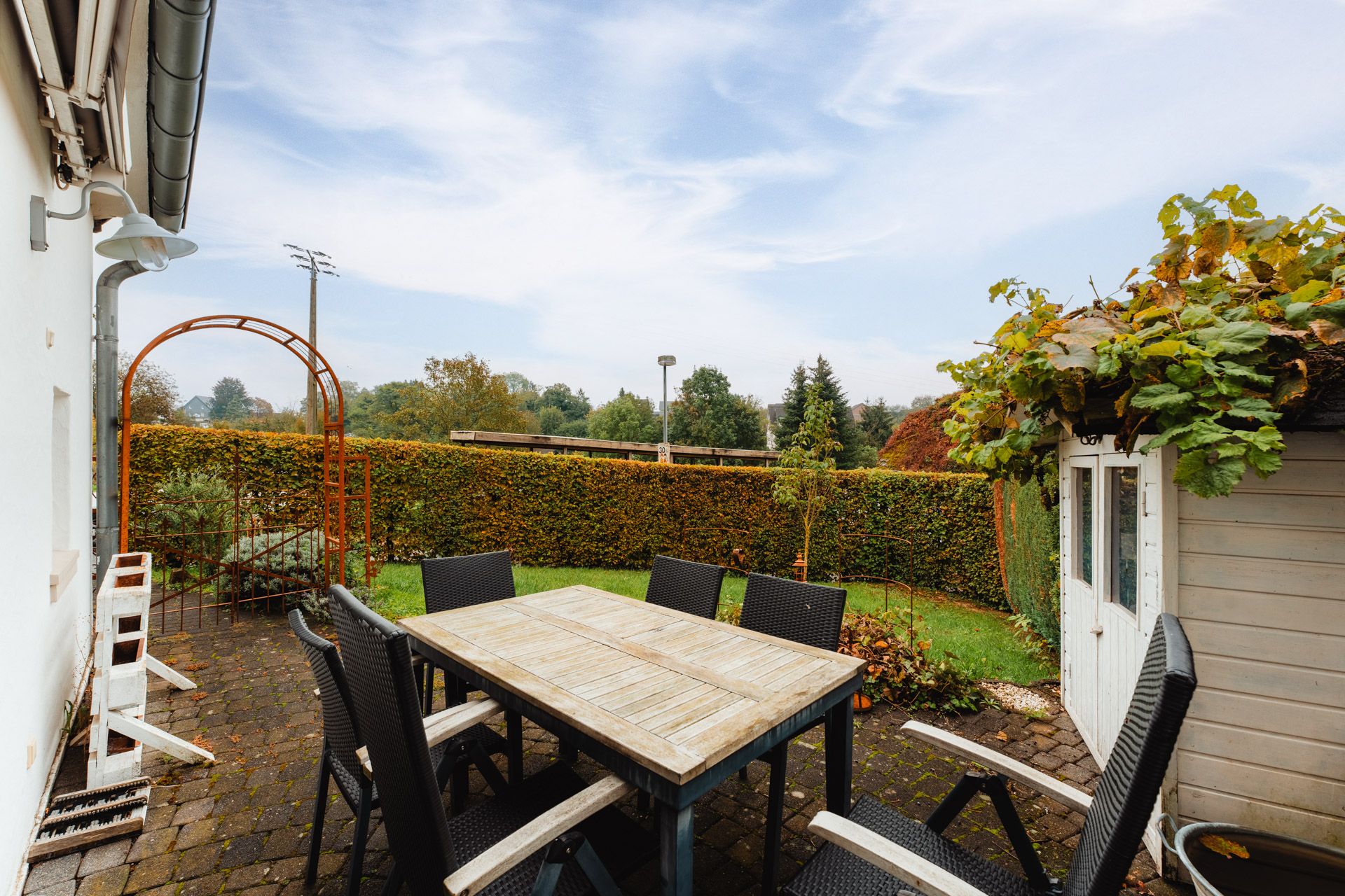 Gepflasterte Terrasse mit Holztisch, sechs Stühlen und Gartenhaus im grünen Garten.