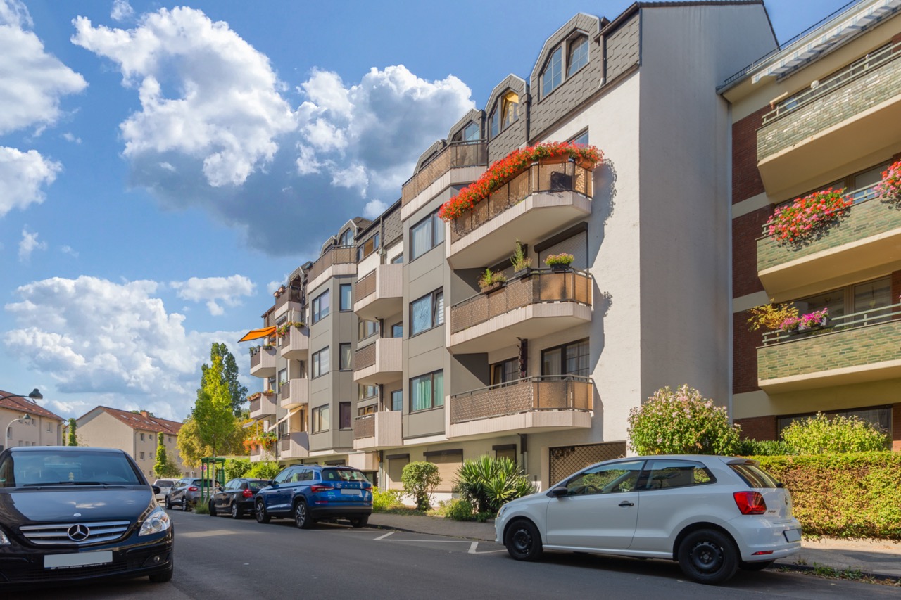 Mehrfamilienhaus mit Balkonen und Blumen, parkende Autos davor, blauer Himmel mit Wolken.