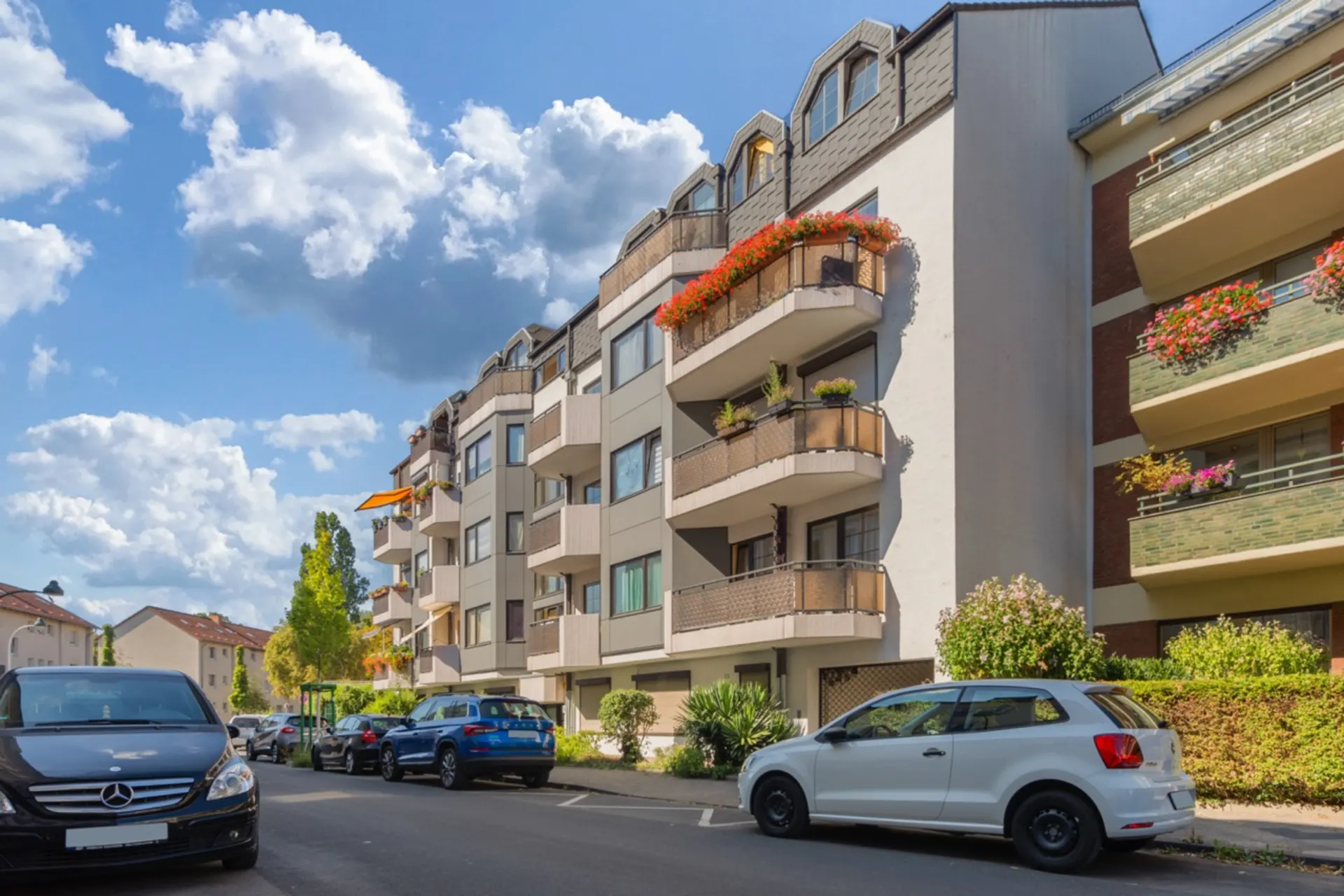 Mehrfamilienhaus mit Balkonen und Blumen, parkende Autos davor, blauer Himmel mit Wolken.