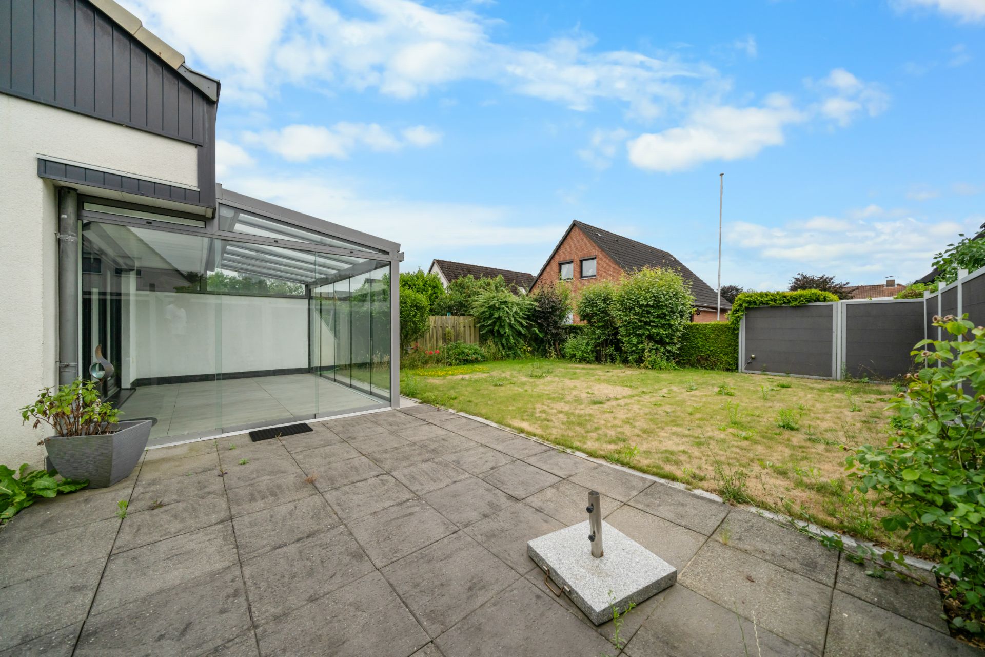 Gepflasterte Terrasse mit Glasüberdachung und angrenzendem Rasen im Garten.