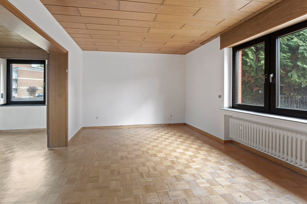 Leerer Raum mit Parkettboden, Holzdecke und großen Fenstern mit Blick ins Grüne.
