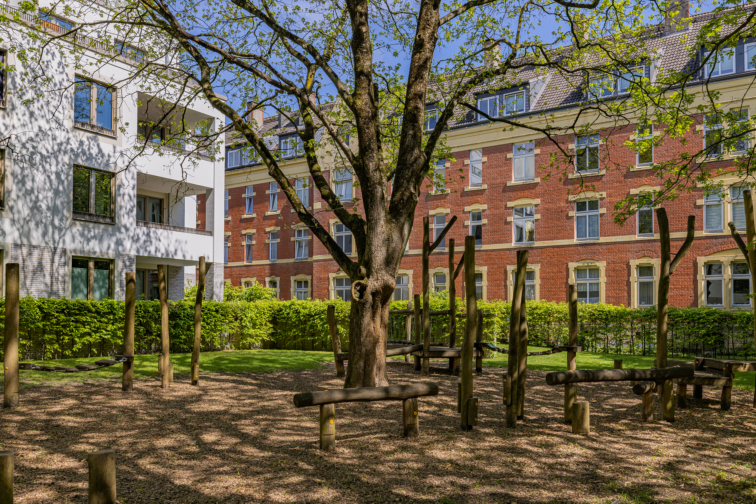 Spielplatz mit Holzbalken und großem Baum im Innenhof vor Wohngebäuden.