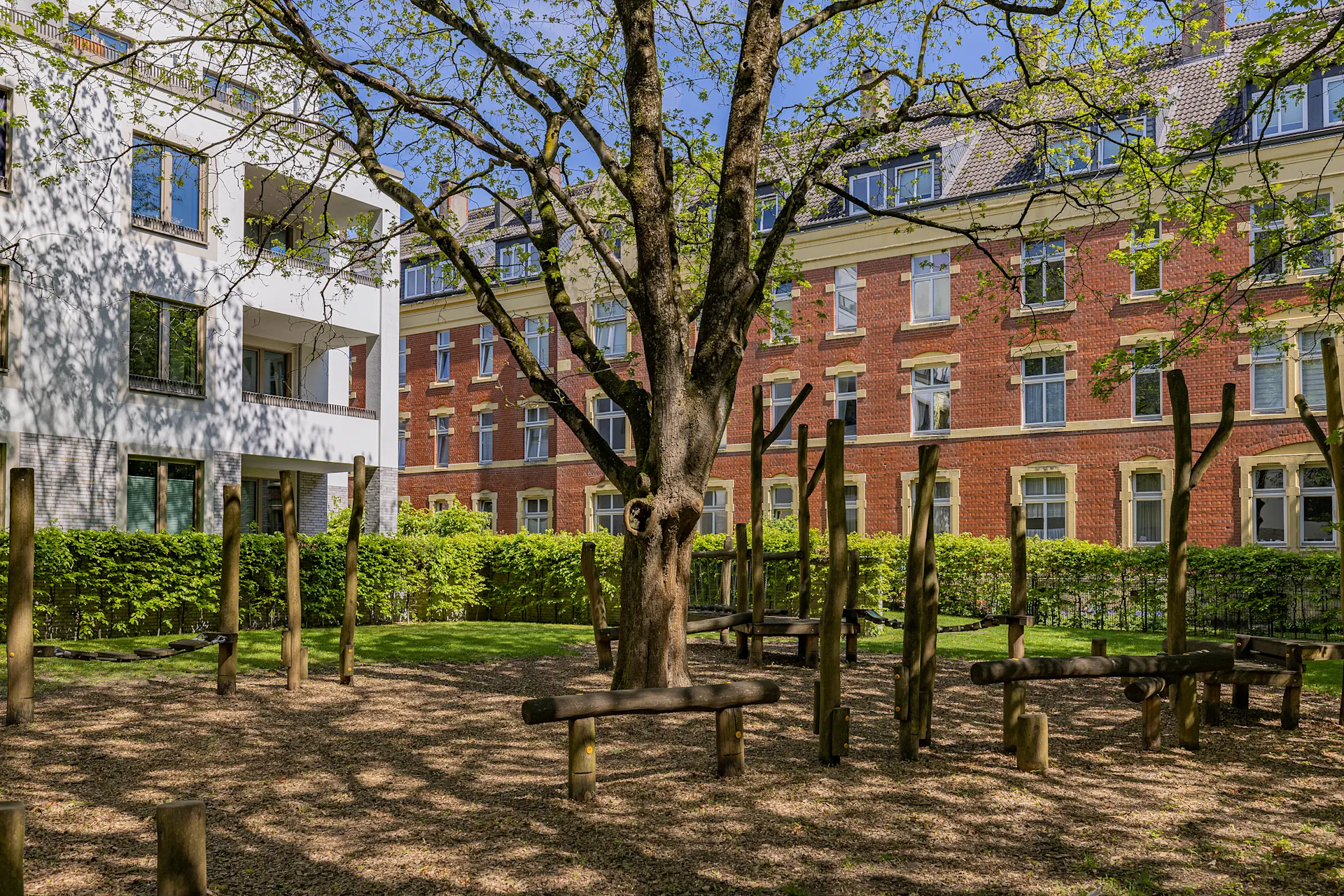Spielplatz mit Holzbalken und großem Baum im Innenhof vor Wohngebäuden.