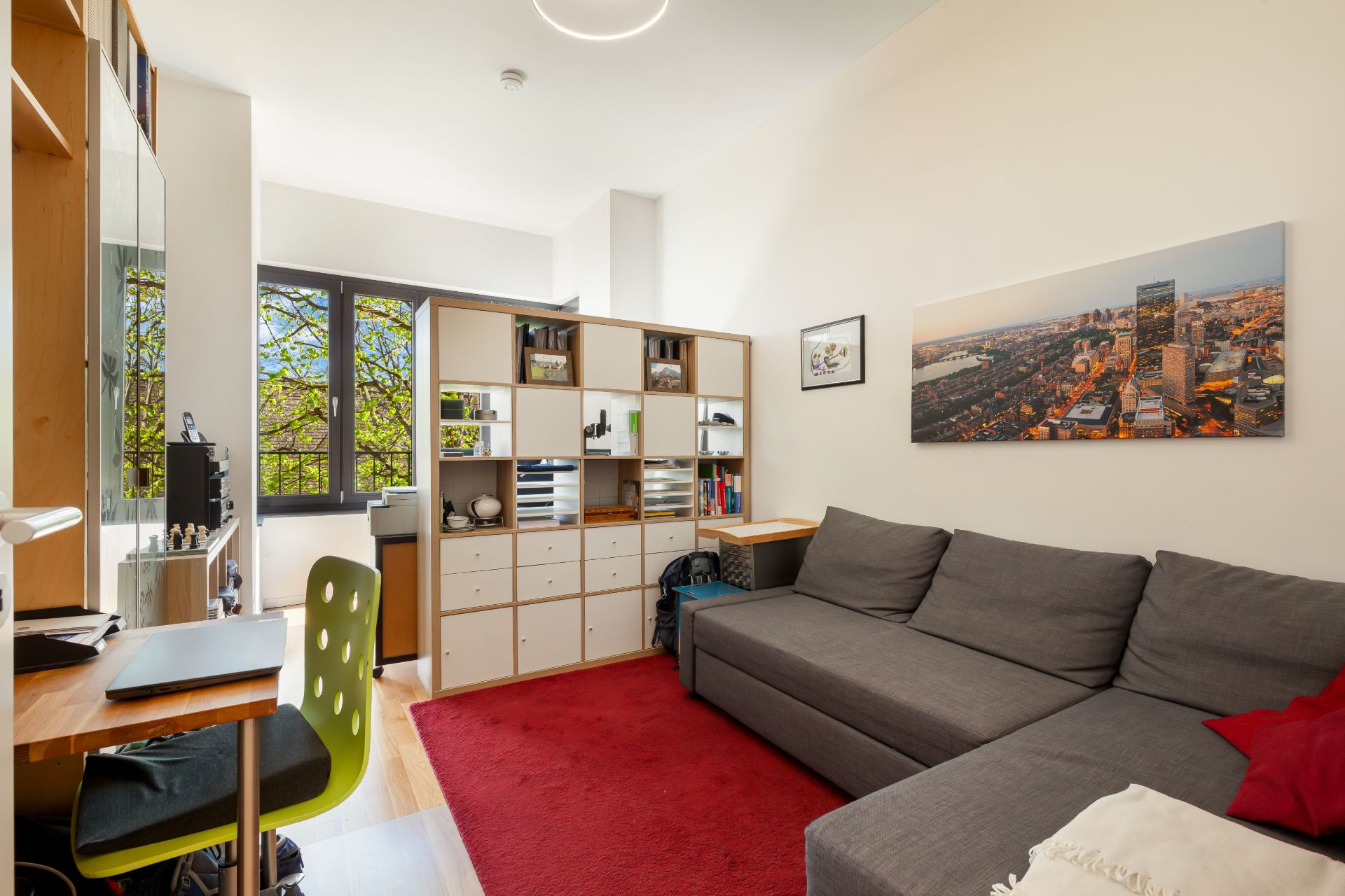 Helles Wohnzimmer mit grauem Sofa, rotem Teppich, Regalen und Schreibtisch am Fenster.