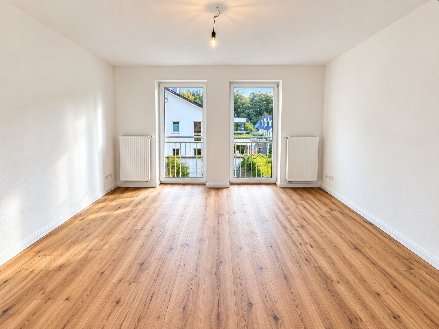 Helles, leeres Zimmer mit Holzboden, weißen Wänden und zwei Fenstertüren mit Blick nach draußen.