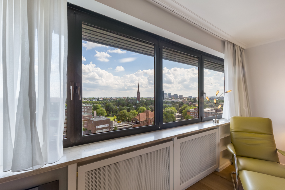 Großes Fenster mit Blick auf Stadt und grüne Bäume, daneben ein grüner Sessel und weiße Vorhänge.