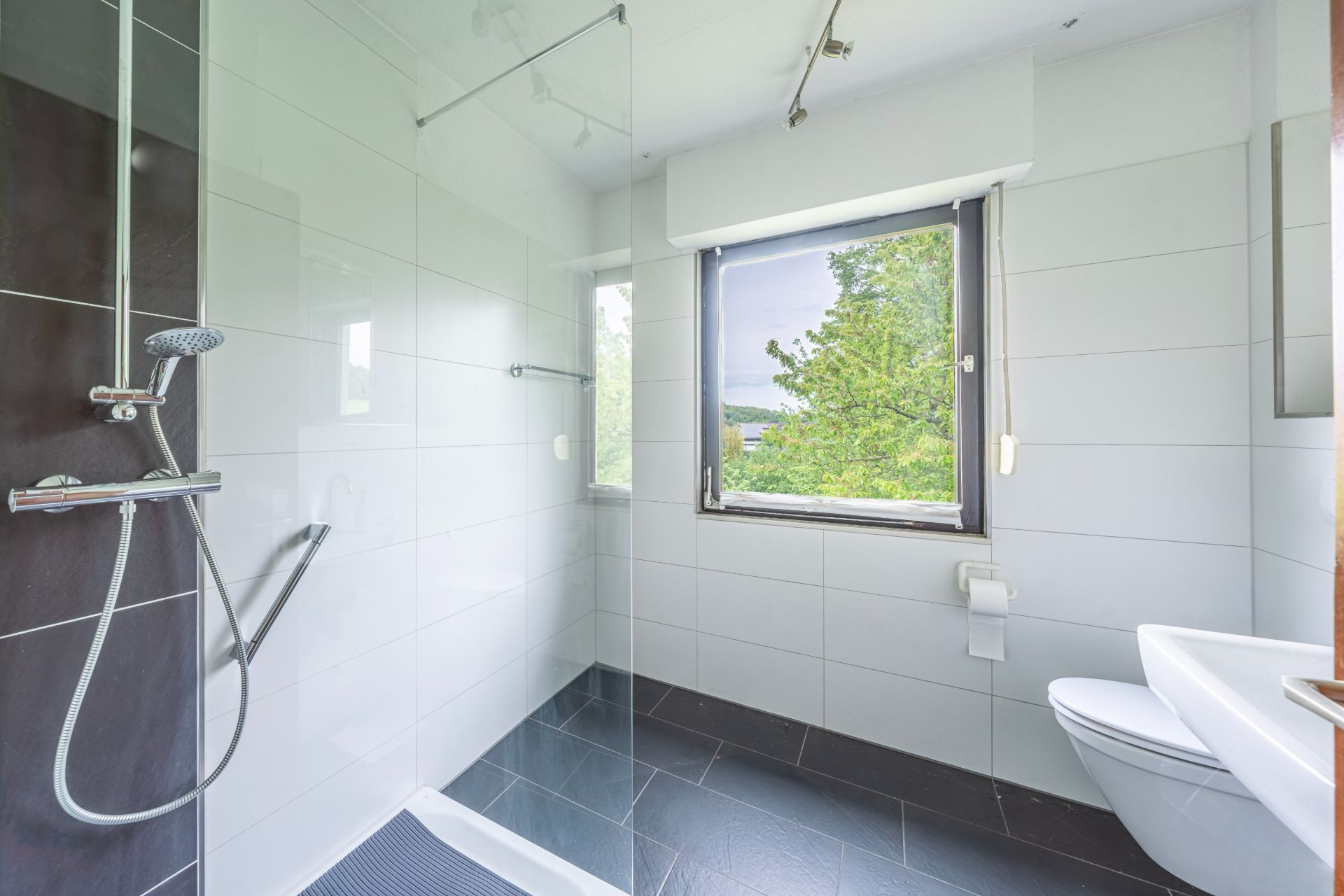 Modernes Badezimmer mit Dusche, Fenster, WC und weißen Fliesen.