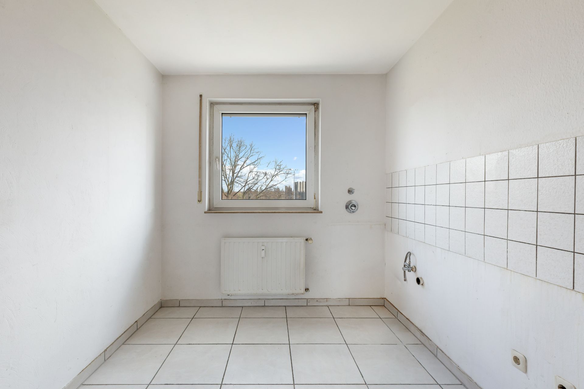 Heller, leerer Raum mit Fliesenboden, weißer Wand, Fenster und Heizkörper.