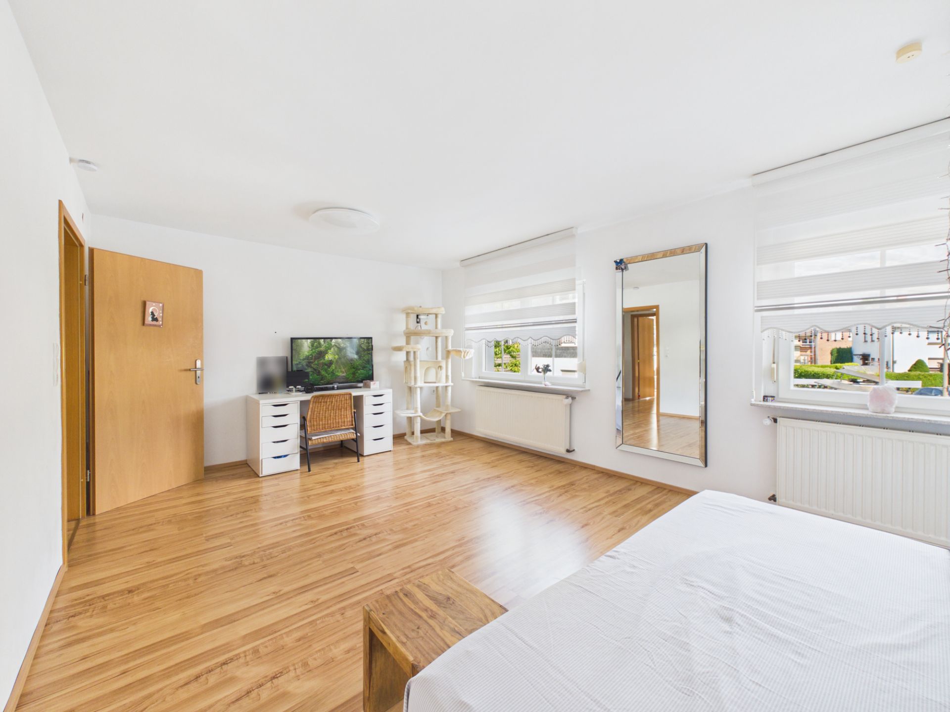 Helles Schlafzimmer mit Holzfußboden, Bett, Schreibtisch, Spiegel und großen Fenstern.