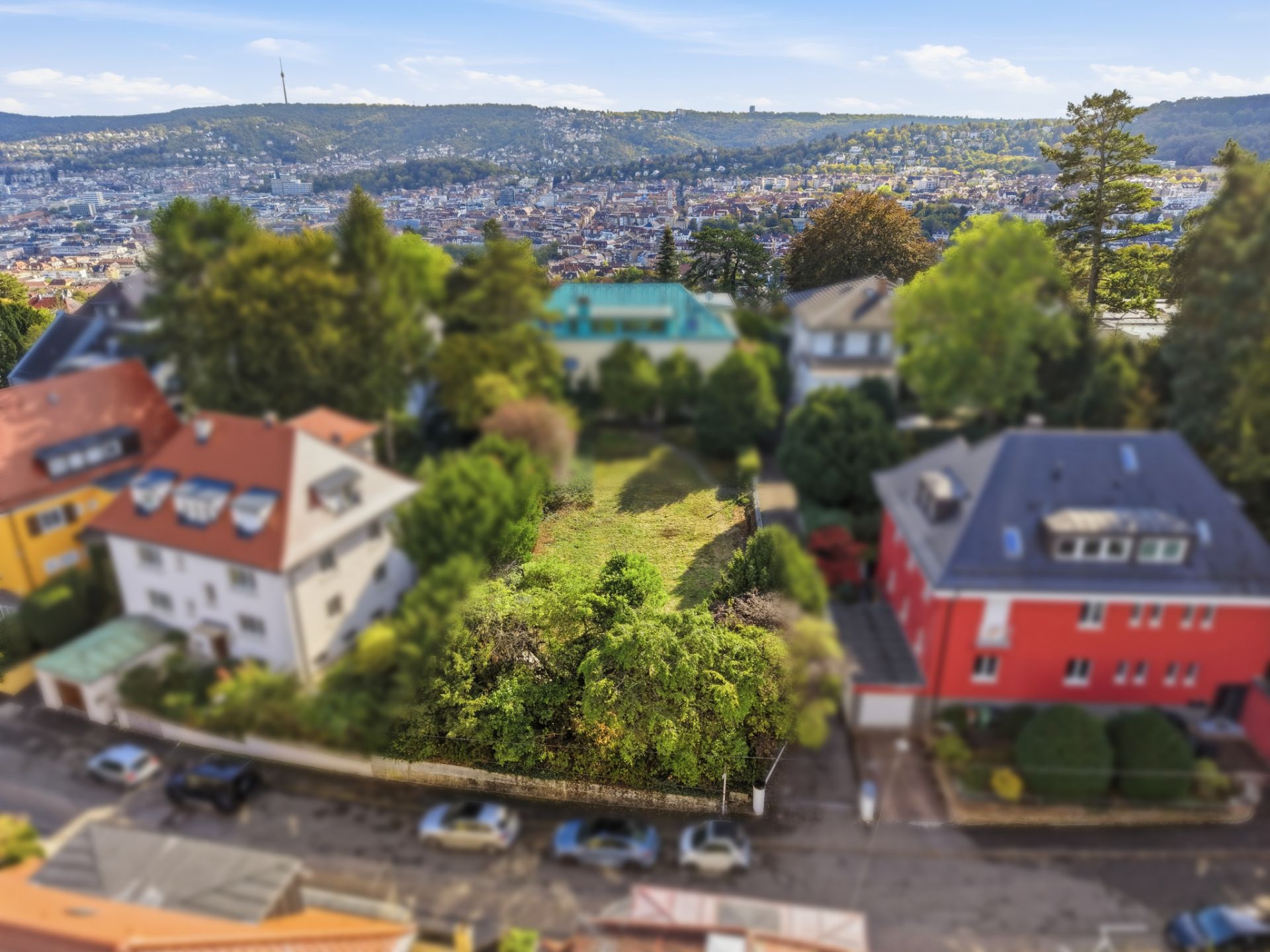 Blick auf ein grünes Grundstück zwischen Wohnhäusern in einer Stadt mit Hügeln im Hintergrund.