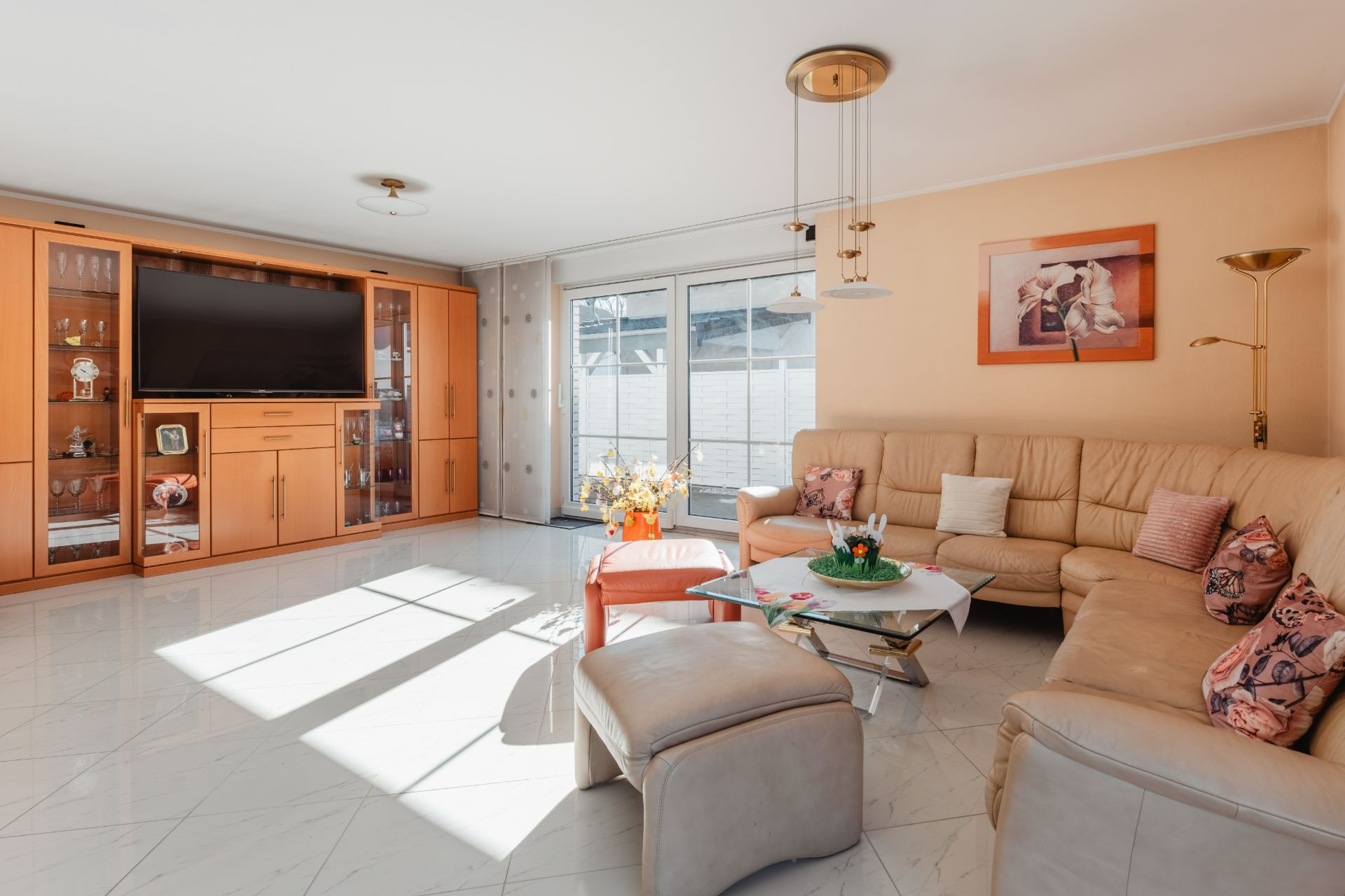 Helles Wohnzimmer mit Ecksofa, Glastisch, TV-Schrank und großen Fenstern.