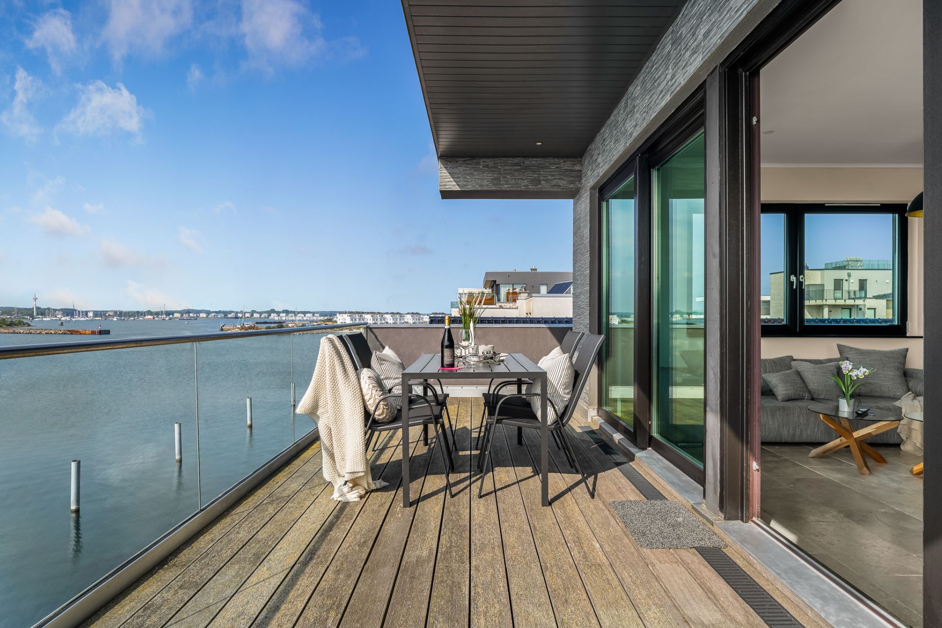 Balkon mit Holzboden, Tisch und Stühlen, Blick auf Wasser und moderne Gebäude.