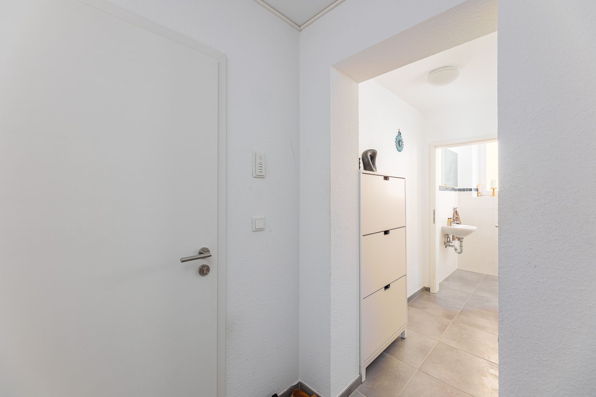 Heller Flur mit weißer Tür, Schuhschrank und Blick in ein Badezimmer mit Waschbecken.
