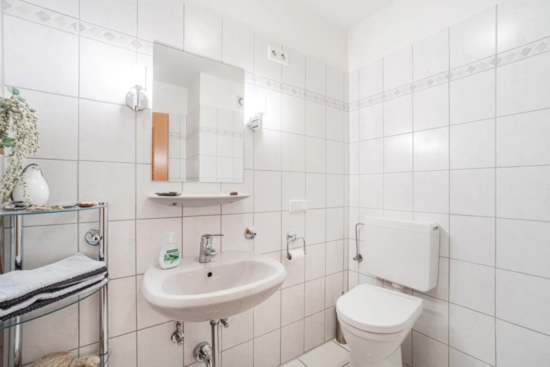 Helles Badezimmer mit Waschbecken, Spiegel, Toilette und Regal mit Handtüchern.