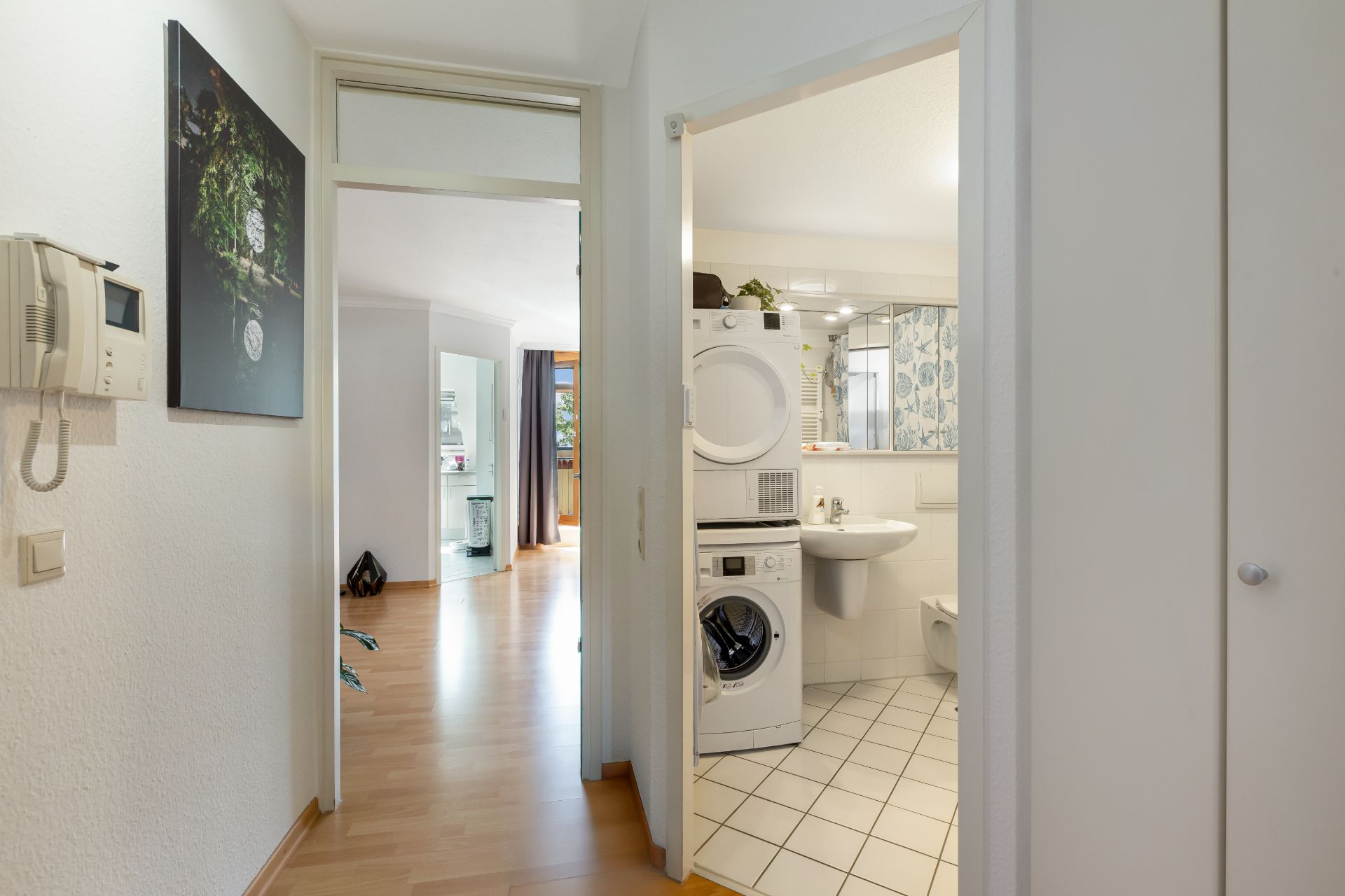 Blick vom Flur auf ein Bad mit Waschmaschine und Trockner, daneben ein Wohnzimmer.