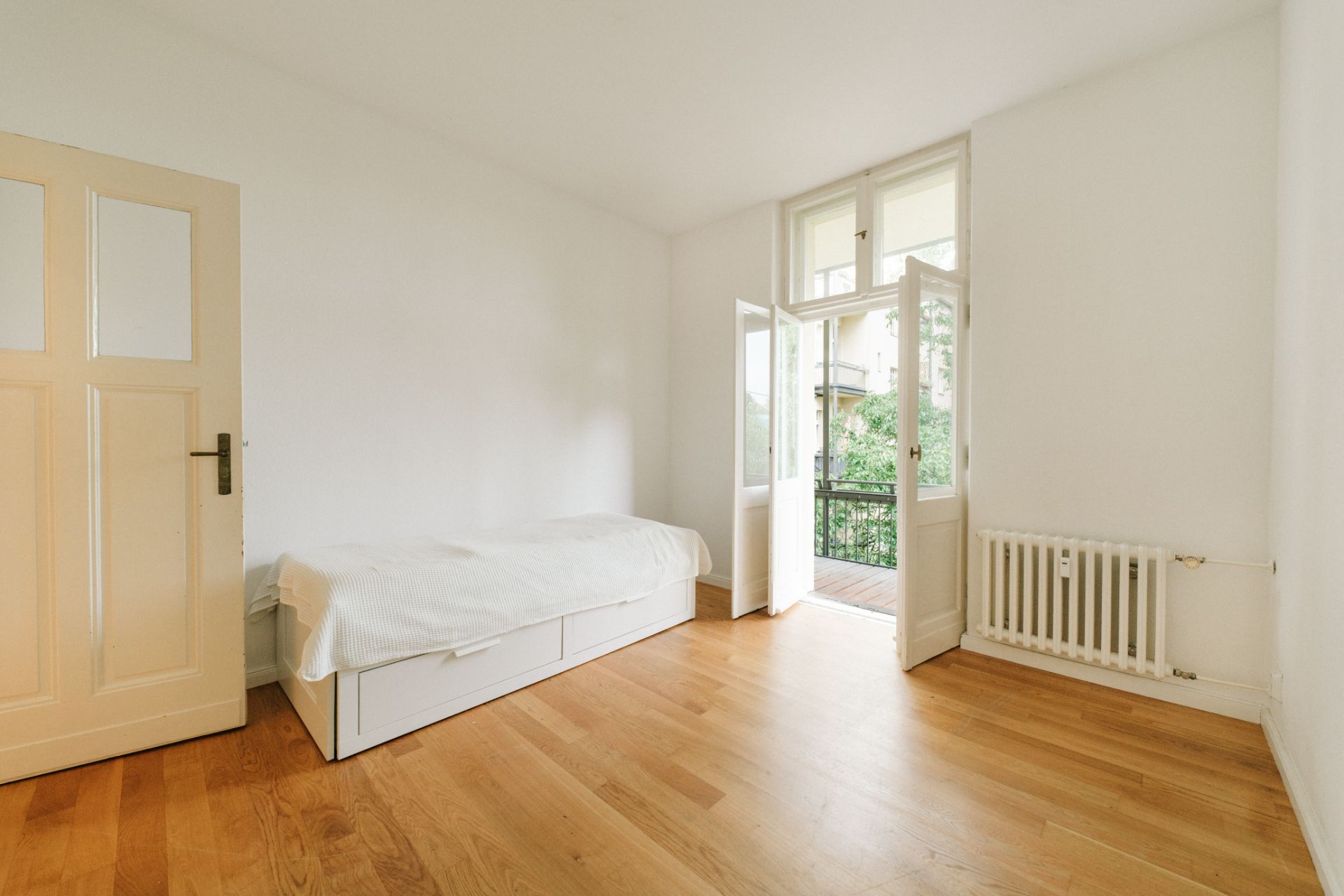 Helles Zimmer mit Holzboden, Einzelbett, Heizung und offenem Zugang zu einem Balkon.