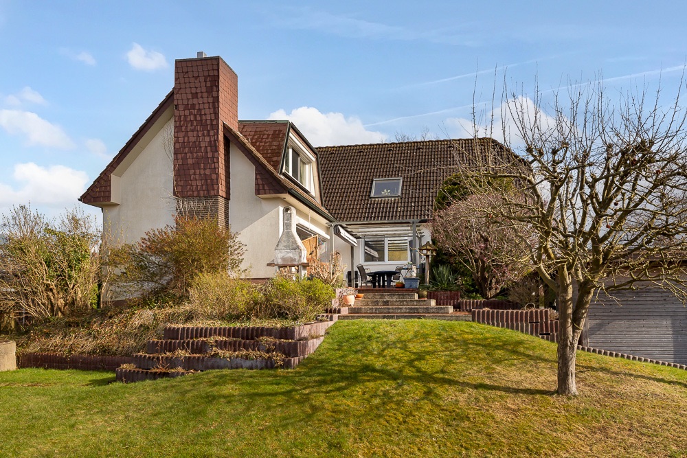 Ein Einfamilienhaus mit Garten, Terrasse und Grill im Vordergrund bei sonnigem Wetter.