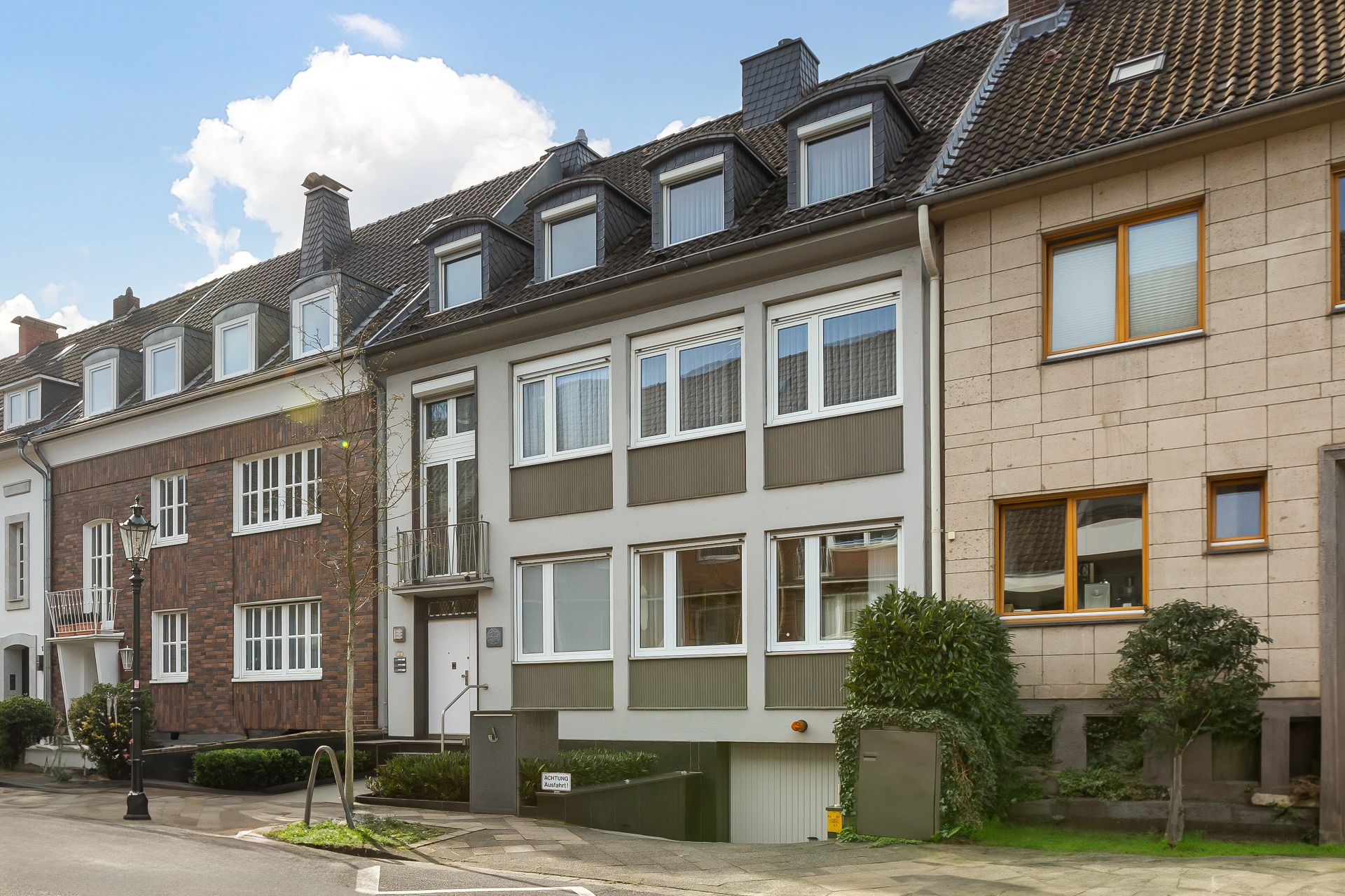 Mehrfamilienhaus mit mehreren Fenstern, Garageneinfahrt und kleinen Bäumen an der Straße.