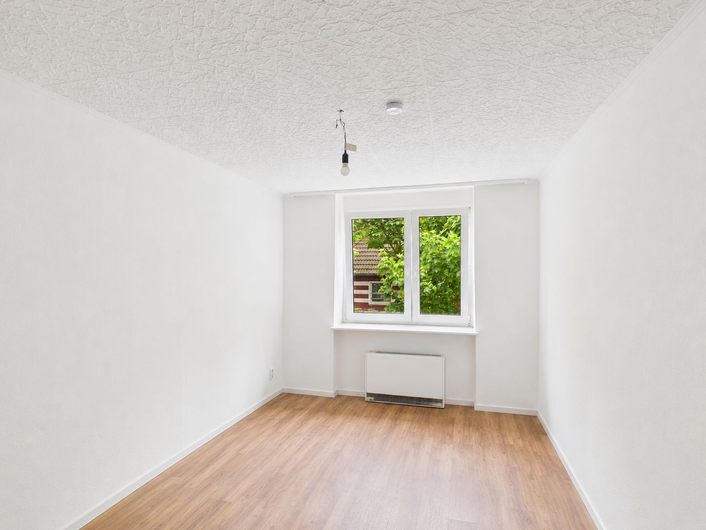 Leeres Zimmer mit weißen Wänden, Holzfußboden und Fenster mit Blick auf Bäume.