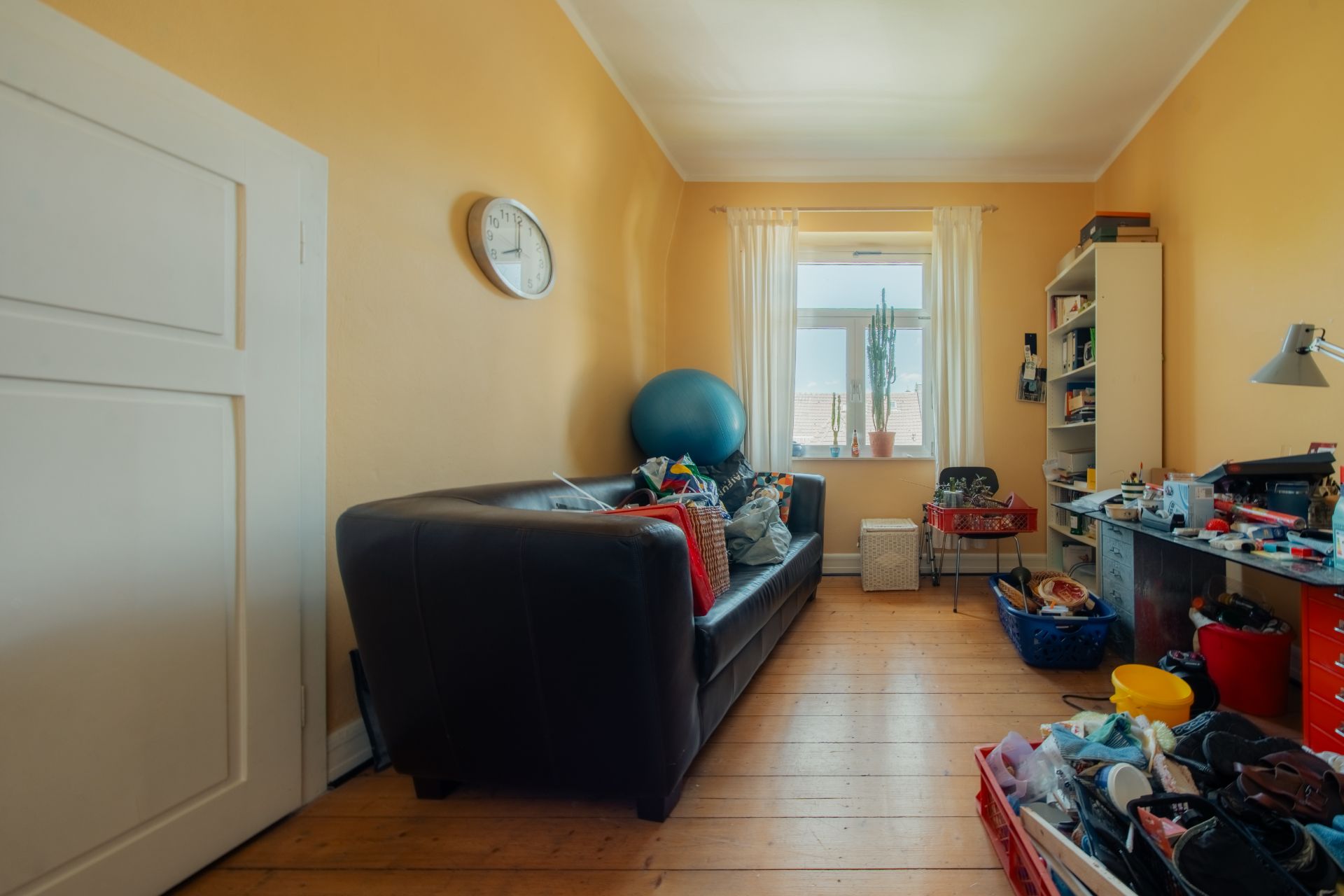 Kleines Zimmer mit braunem Sofa, Schreibtisch, Wandregal und Fenster mit Vorhängen.