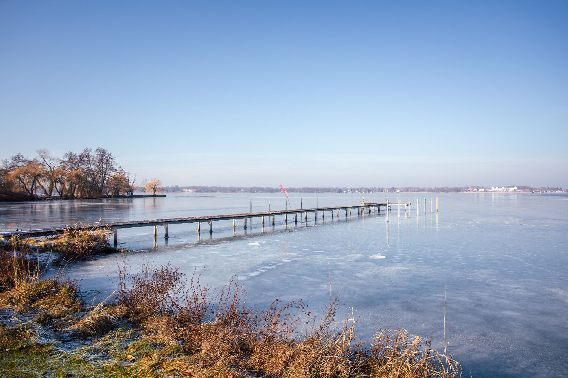 Eisiger See mit Steg und winterlicher Vegetation am Ufer bei klarem Himmel.