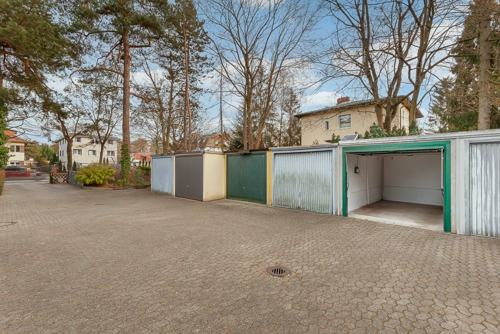 Mehrere Garagen mit geöffnetem Tor und gepflastertem Vorplatz zwischen Bäumen und Wohnhäusern.