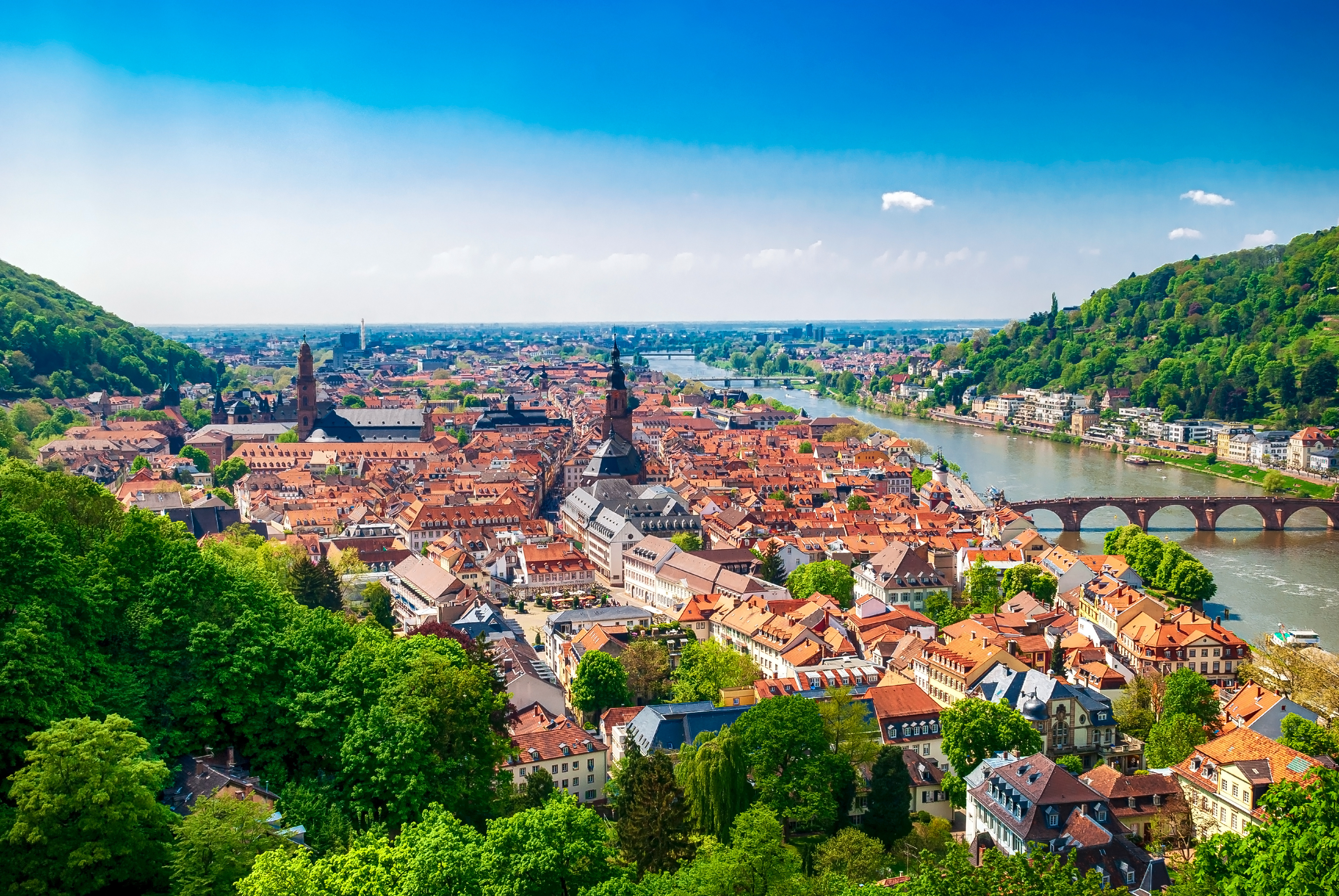 Blick auf eine Stadt mit roten Dächern, Fluss und Brücke, umgeben von grünen Hügeln.