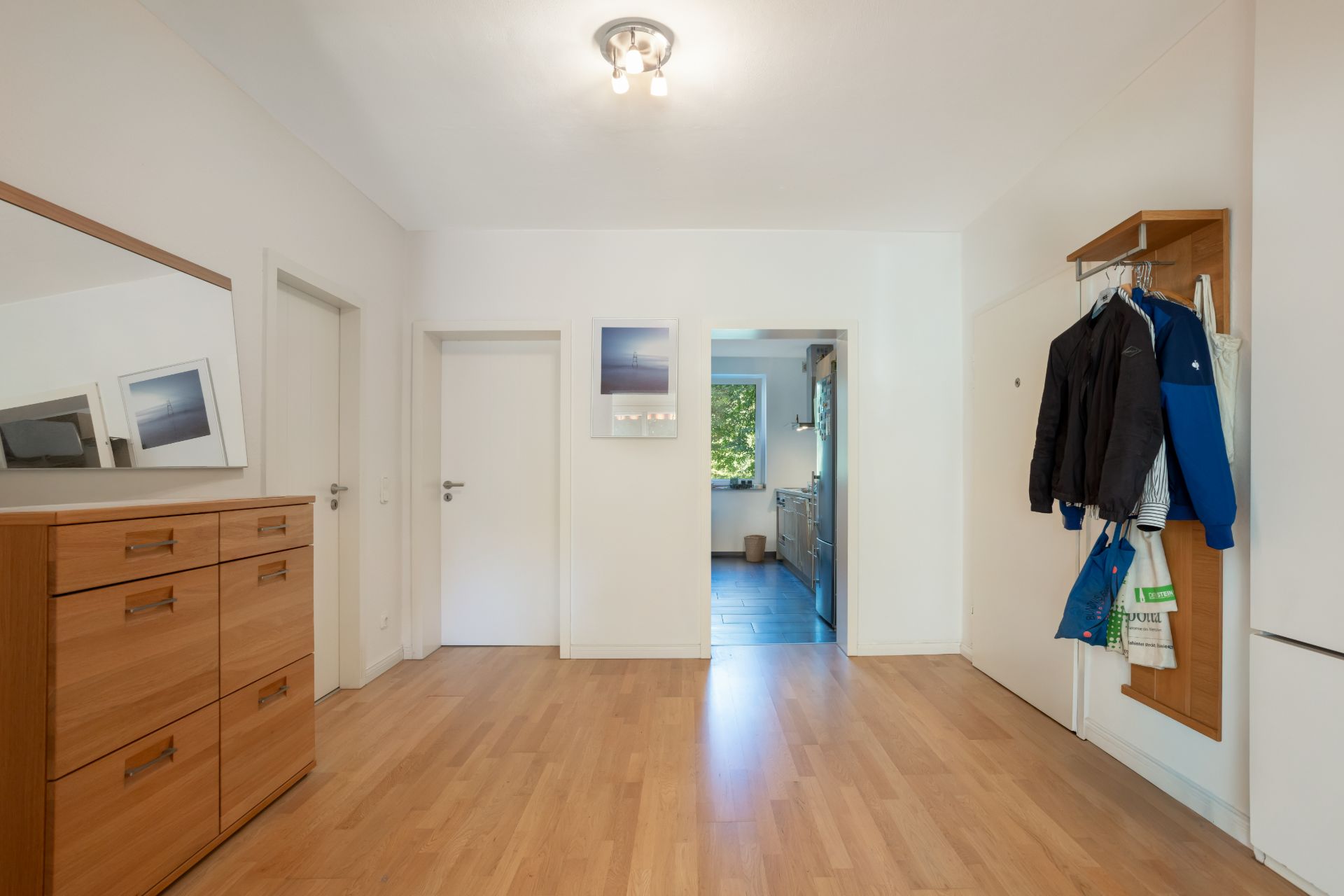 Heller Flur mit Holzkommode, Garderobe, Türen und Blick in eine Küche.
