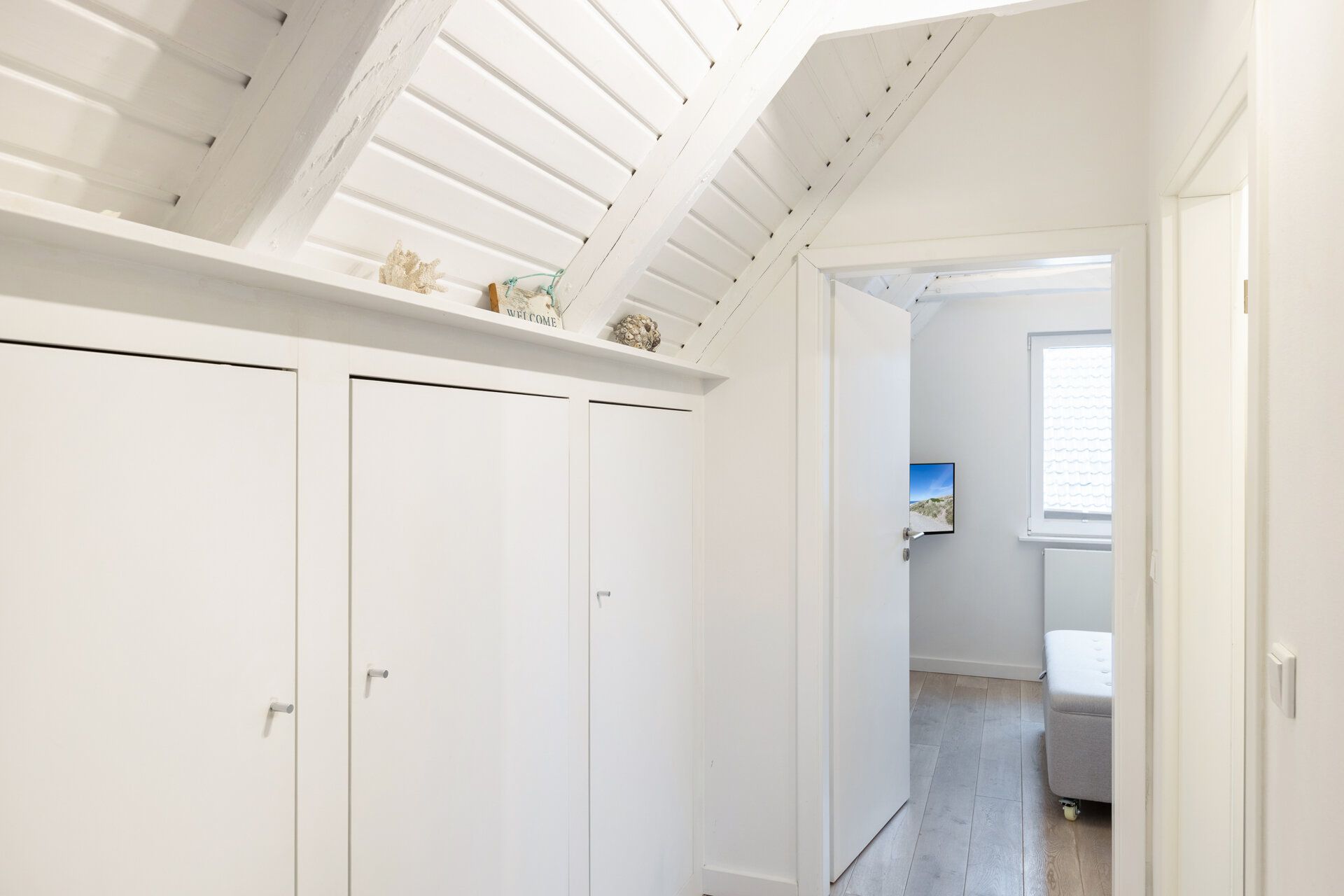 Heller Flur mit weißen Einbauschränken und schräger Decke, Tür zu einem Zimmer mit Fenster.
