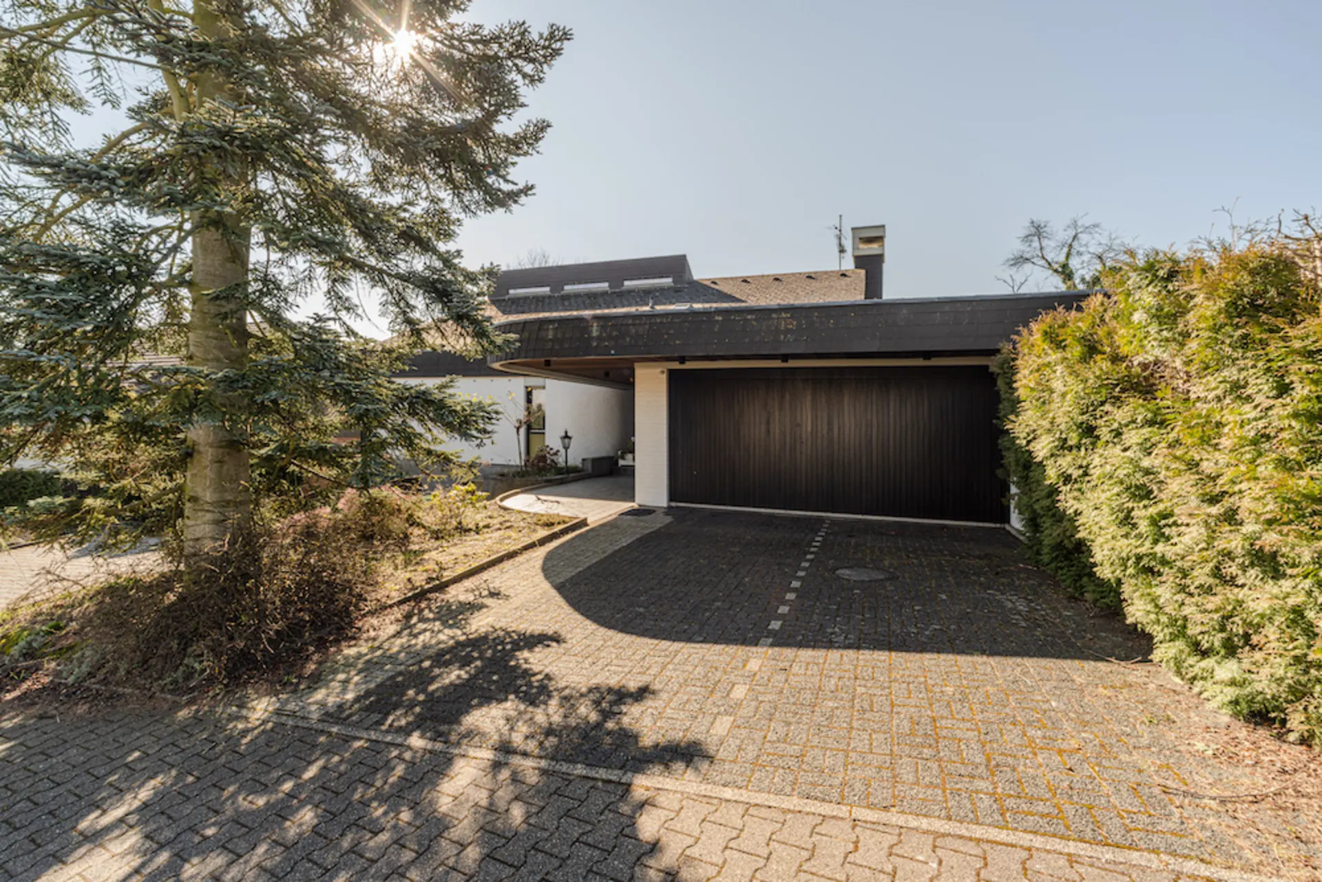 Einfamilienhaus mit dunkler Garage, Auffahrt, Baum und Hecke bei Sonnenschein.